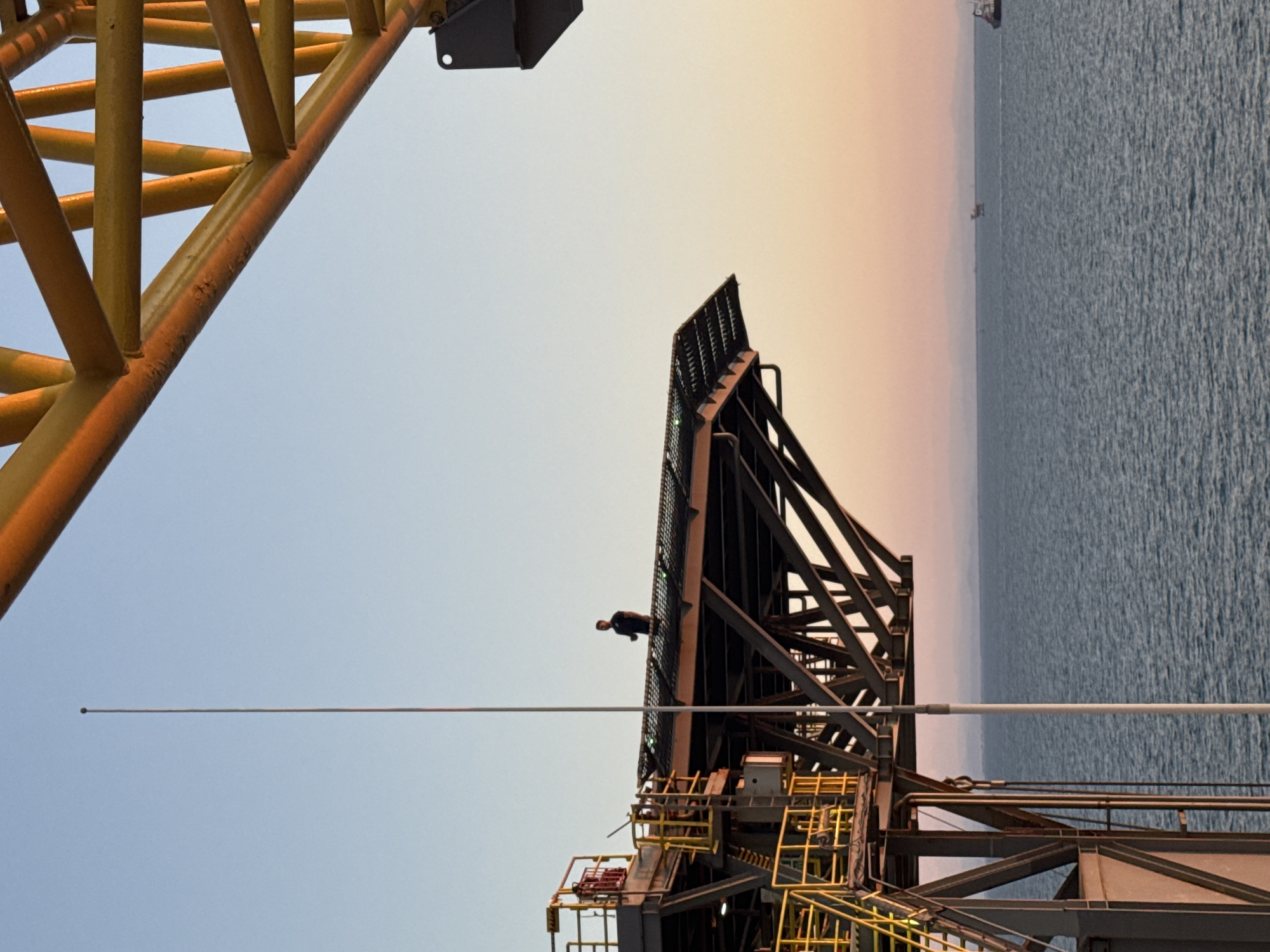 The image depicts an outdoor scene, likely at sea given the presence of water and distant vessels. In the foreground, a person stands on a platform of what appears to be an industrial structure, possibly an oil rig or a large maritime platform, with a complex metal framework. The platform is elevated above the sea, and a tall, thin mast extends upwards into the clear sky. The sky transitions from a pale blue near the horizon to a soft orange and pink at the top, indicating either sunrise or sunset. A large, yellow, geometric structure, also part of a larger industrial installation, dominates the upper right portion of the frame. In the distance, several other vessels or structures can be seen on the water. The overall impression is one of an industrial setting in a marine environment during twilight.