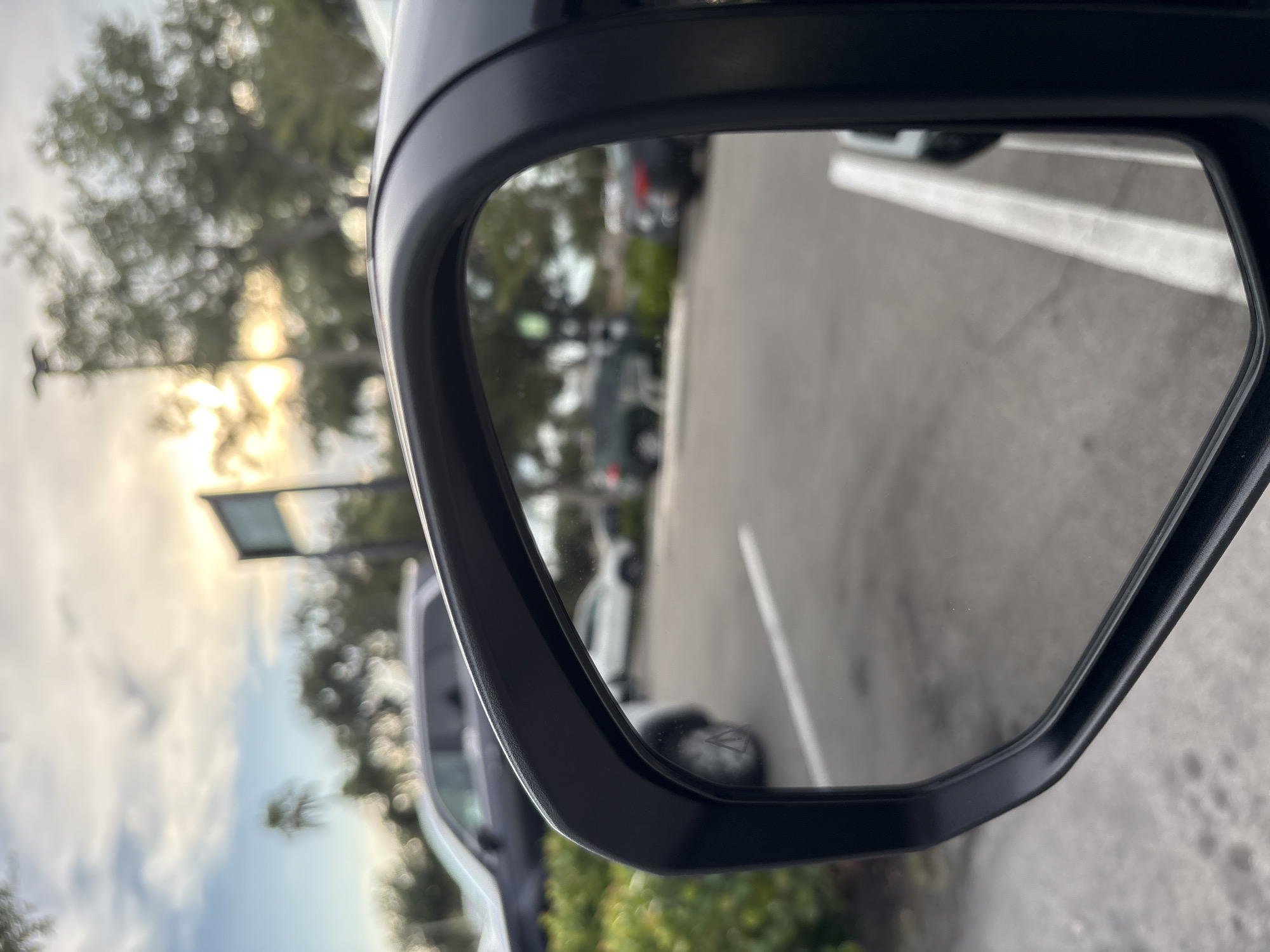 The image shows a rear-view mirror of a car parked in a lot, with a view of other cars and trees reflected in the mirror. The sky is cloudy with sunlight breaking through. The scene appears to be in South Venice, United States of America. There are several cars visible in the reflection, including a dark SUV, a white SUV, and a dark sedan. The environment is a parking lot with some greenery and trees in the background. No people are visible in the image.
