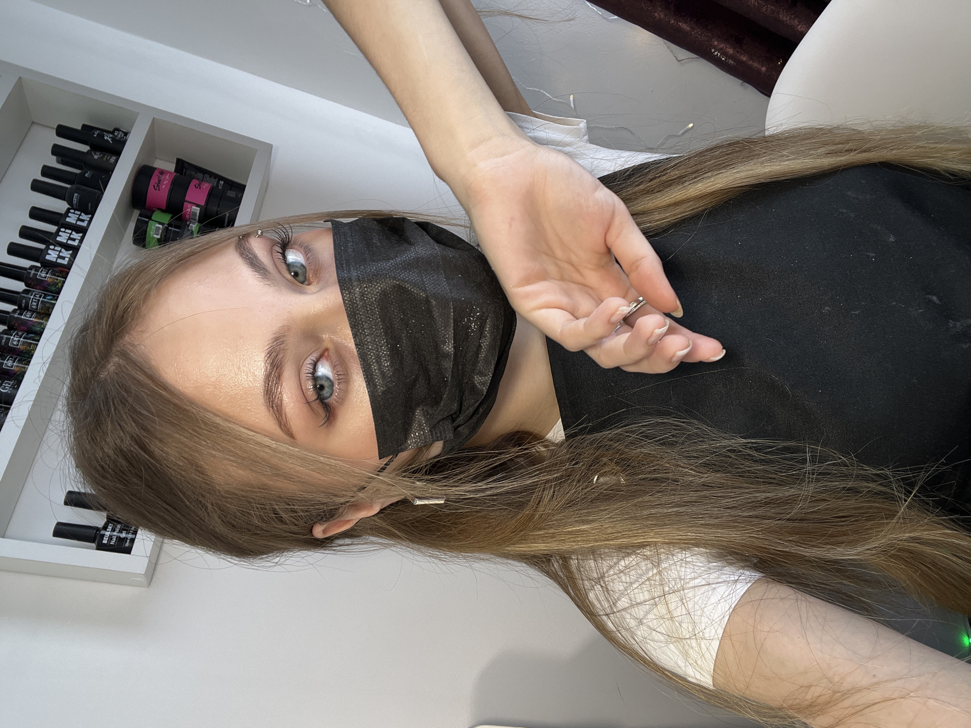 A young person wearing a black face mask is lying down. Part of the right side of their face is visible, showing a piercing on their eyebrow, blue eyes, and blond hair. Their hair is long and straight. Someone is holding their hand near the face of the person lying down. A shelf with numerous nail polish bottles is visible in the background on the left side of the image. A white chair is partially visible behind the person. Based on the nail polish supplies, this scene could take place in a nail salon. There is no definitive evidence the scene is in Фоки, Russia.
