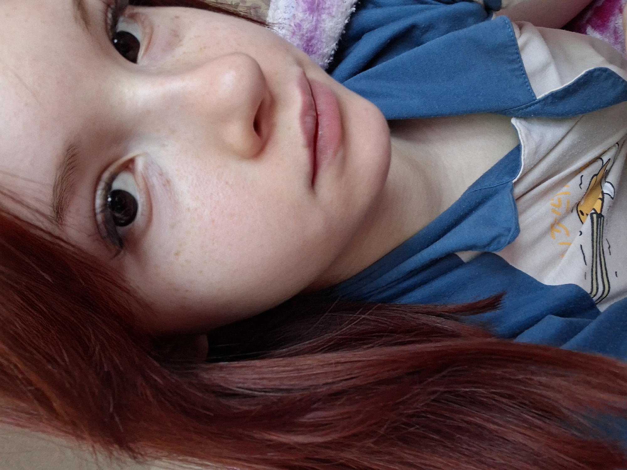 A close-up, angled selfie shows a young, fair-skinned individual with red hair and freckles. They have dark eyes and are looking upward towards the camera. The person is wearing a blue, collared shirt over a lighter colored top with a graphic design. A soft, possibly hooded, garment with a light purple and white pattern is visible behind the person's head. The background is indistinct and out of focus. It is not possible to determine the location in the city of Chaikovsky, Russia from this image.