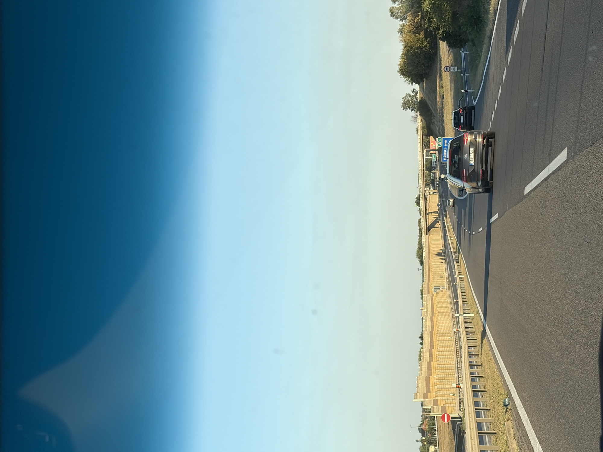 The image shows a view from a vehicle traveling on a highway in Križovany nad Dudváhom, Slovakia. The highway has multiple lanes and is bordered by a guardrail and a sound barrier. Several cars are visible on the highway, including a dark gray station wagon in the foreground. In the distance, a road sign indicates "VLČKOVCE - SOVĚR". The sky is clear and blue, with the sun casting long shadows. The surrounding environment appears to be rural, with trees and vegetation visible on the sides of the highway.