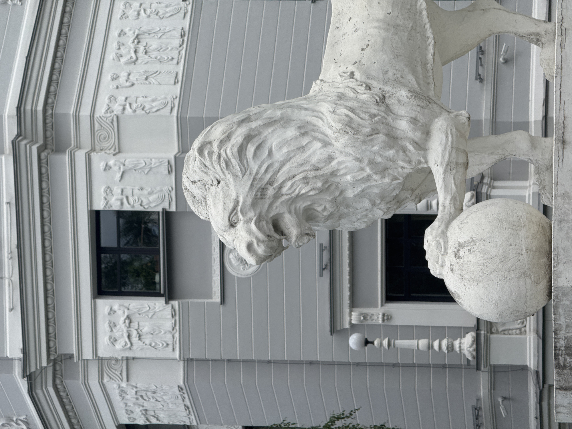A weathered, white stone sculpture of a lion is positioned in the foreground, its mouth open in a roar, with one paw resting on a large sphere. The lion is situated in front of an imposing, grey, classical-style building adorned with white decorative relief carvings of figures. The building features multiple windows with dark frames and horizontal detailing. A lamppost with a white globe stands to the left of the lion sculpture. The image appears to be taken outdoors during the daytime, with overcast lighting. The location is identified as Yekaterinburg, Russia.