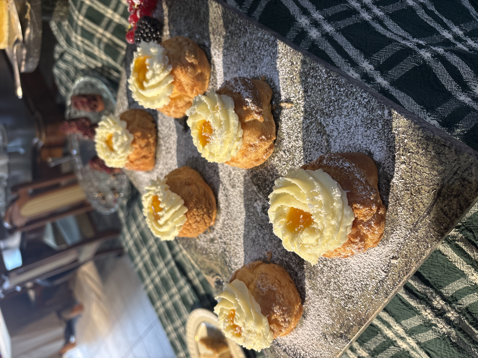Six cream puffs, dusted with powdered sugar and topped with white frosting and orange jam, are arranged on a dark stone platter. The platter sits on a green and white checkered tablecloth. In the background, a wooden table and chairs are visible, along with a blurred image of what appears to be a plate of pastries and a bowl of berries. The scene is illuminated by sunlight, casting shadows. No specific location in Riga, Latvia is identifiable from the image.
