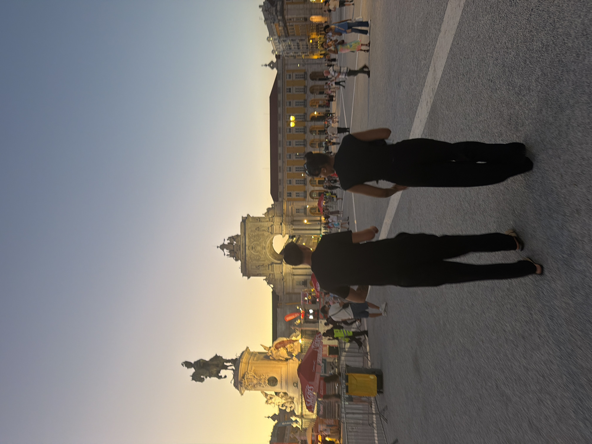 The image captures a view of Praça do Comércio in Lisbon, Portugal, at dusk. The sky is a gradient of light blue to a warm yellow-orange near the horizon. In the background, the Arco da Rua Augusta stands prominently, illuminated by streetlights. To the left, a grand building with a colonnaded facade lines the square. A bronze equestrian statue is visible on a pedestal, also lit.
In the foreground, two people, a man and a woman, are walking away from the camera towards the arch. Both are dressed in dark clothing. The man wears a black t-shirt and black pants. The woman wears a black sleeveless top and wide-legged black pants. Several other people are scattered throughout the square, some walking, some standing. Various structures, including tents and barriers, are set up in the middle ground, suggesting an event or temporary setup in the plaza. The ground is paved with cobblestones, and a white line marks a path on the pavement. The overall atmosphere is that of a public square in the early evening, with a mix of architectural grandeur and pedestrian activity.