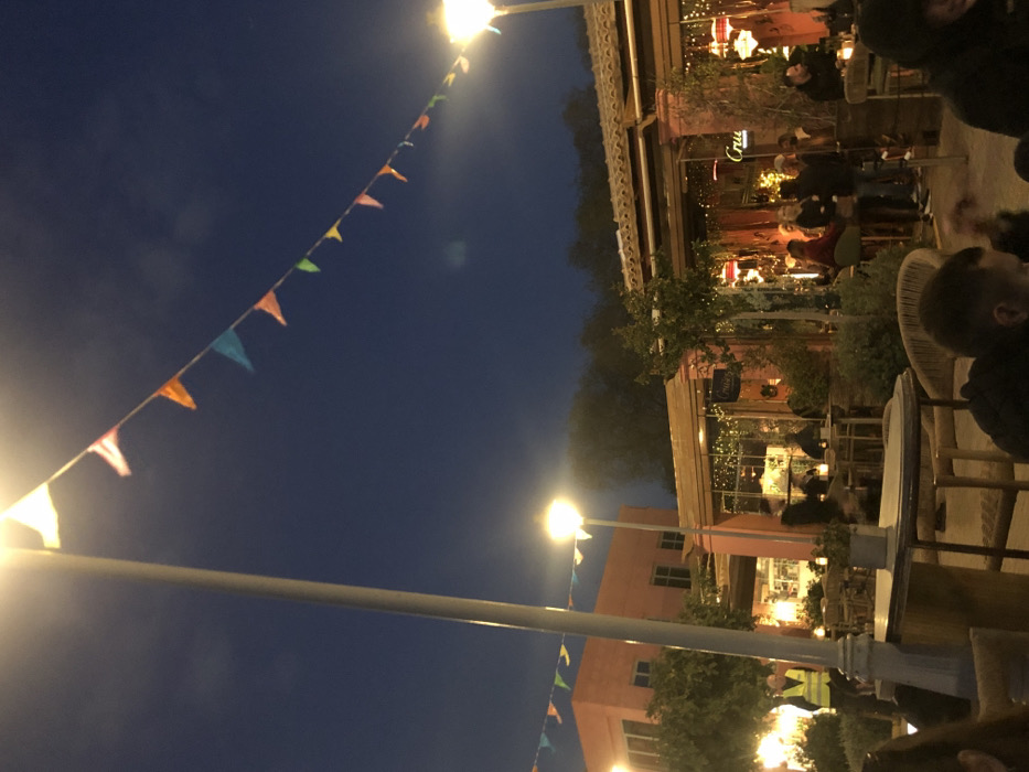 A string of colorful triangular flags stretches across a dark blue night sky, illuminated by two streetlights. The flags are strung between a white pole on the left and a building on the right. The building is a two-story structure with a pink facade and a series of windows. The building is partially obscured by a leafy tree. The scene is located in Palaio Faliro, Greece, at the coordinates 37.929 latitude and 23.685 longitude. The building is a restaurant and has a sign that says "Cru" on the upper level. People are sitting at tables and enjoying the evening atmosphere. There are multiple lights in the area that illuminate the scene. The light from the streetlights and building lights illuminate the space, providing a warm and inviting ambiance. A white table with a white top and a white base stands on the ground, just under the streetlight on the right.