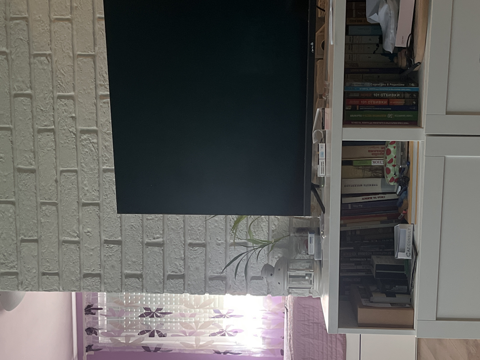 A close-up shot shows a section of a room, featuring a white brick wall, a computer monitor, and a white bookshelf filled with books. The books have titles in Bulgarian, some of which include "101 ОТБИВКИ" and "СИЛА ЗА ЖИВОТ." There's also a small plant near the monitor. A patterned curtain is visible below the wall and monitor.