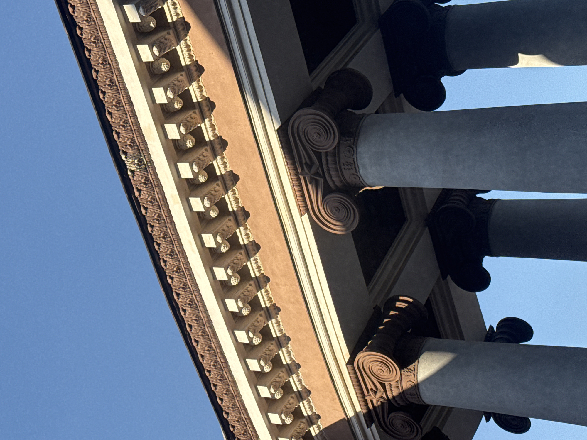 This media file captures an upward-looking perspective of architectural elements, likely from a classical-style building in Kryvyi Rih, Ukraine. The primary subjects are large, grey columns with ornate capitals featuring volutes and acanthus leaves. These columns support an overhang or roof structure, the underside of which is visible and adorned with decorative moldings.
The scene is dominated by the interplay of light and shadow, suggesting it was taken during daylight hours. The clear, bright blue sky indicates fair weather. Sunlight casts strong shadows, highlighting the textured surfaces of the architectural details. The angle of the shot emphasizes the grandeur and scale of the construction.
There is no visible text in the image. The overall impression is one of architectural detail and the play of light on a clear day. The specific location within Kryvyi Rih cannot be identified solely from the visual information. The image does not depict any people or activity.