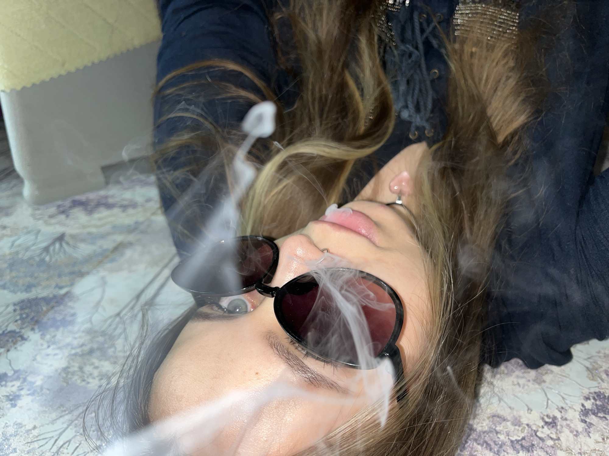 A young woman lies on a patterned surface, likely a bed or floor covering, and exhales smoke. She wears sunglasses and a dark-colored garment, possibly a shirt or jacket, with fringed detailing. Her long hair is a mix of brown and blonde shades. The smoke partially obscures her face and glasses. An off-white object with a textured surface is visible in the periphery. The location in Novopokrovskaya, Russia is not verifiable from the image.