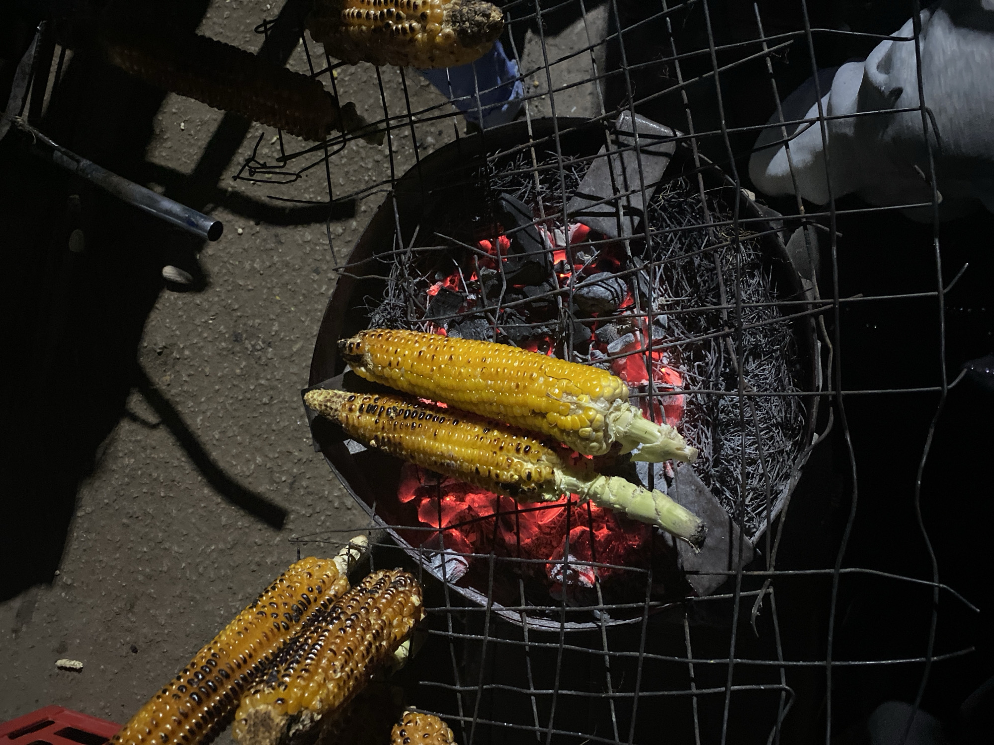 The image shows corn on the cob being grilled in Bukuru, Nigeria. The corn is placed on a wire rack over a charcoal grill with visible red embers. Some corn is partially charred, while others are still a bright yellow color. Additional grilled corn sits to the side. The scene is set at night.