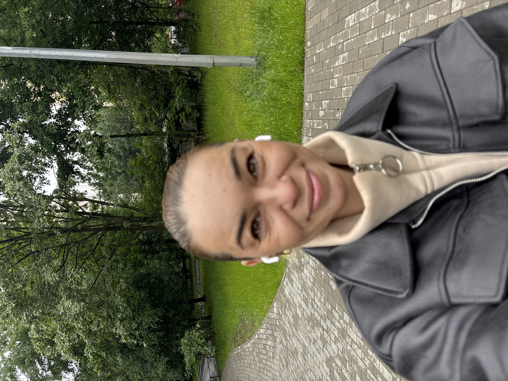 A woman is taking a selfie outdoors in Vsevolozhsk, Russia. She is standing on a paved walkway, possibly a park path, with a grassy area and trees in the background. She is wearing a black leather jacket over a cream-colored hoodie. White earbuds are visible in her ears. The photo is taken from a low angle, with the woman's face in the foreground and slightly out of focus. The trees and foliage suggest it is summer or early autumn. A metal pole is visible to the right of the woman.