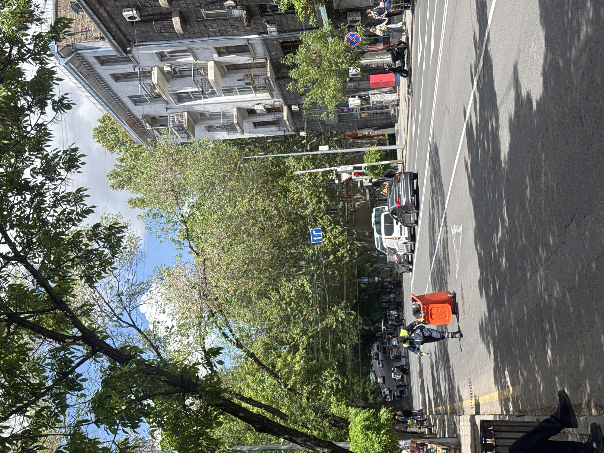 The scene is an urban setting in Yerevan, Armenia. A street runs vertically through the image, lined with buildings and trees. On the right side of the street, a road worker in blue uniform and gloves is bent over with an orange bucket in the center of the road. Parked and moving vehicles are visible on the road. Trees and buildings line the left side of the image with leafy branches and blue sky visible above the trees. A traffic light and street signs are visible amongst the trees. An individual sits on a bench in the lower right corner, their legs and feet visible.