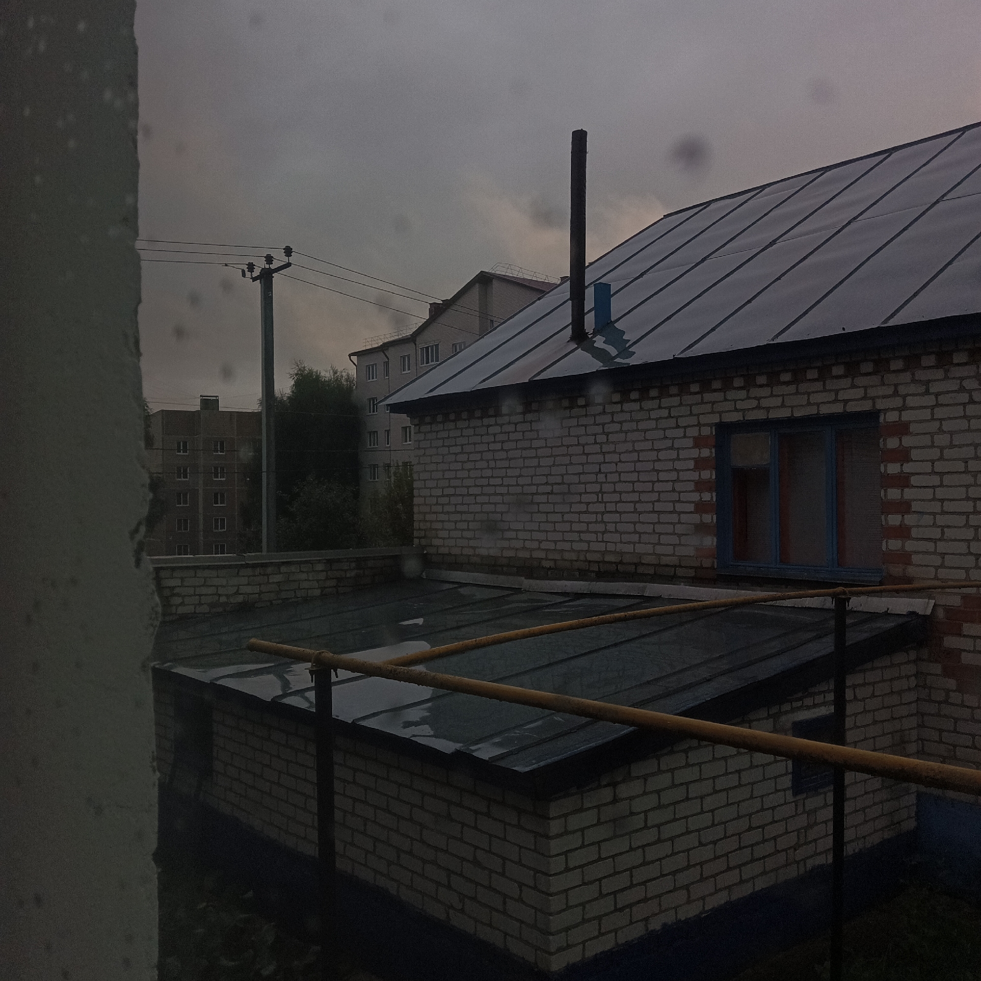 The image depicts an overcast sky over the city of Shumerlya, Russia, seen through a window with water droplets. In the foreground, a textured grey surface occupies the left side, possibly a wall. Adjacent to this is a dark, flat roof, partially obscured by a yellow railing supported by black metal posts. Behind this roof, a brick building with a dark roof and a vertical pipe is visible. Further in the background, a multi-story apartment building with numerous windows stands under the cloudy sky. Another power pole with wires is also present in the scene. The overall impression is one of a gloomy, possibly rainy day.