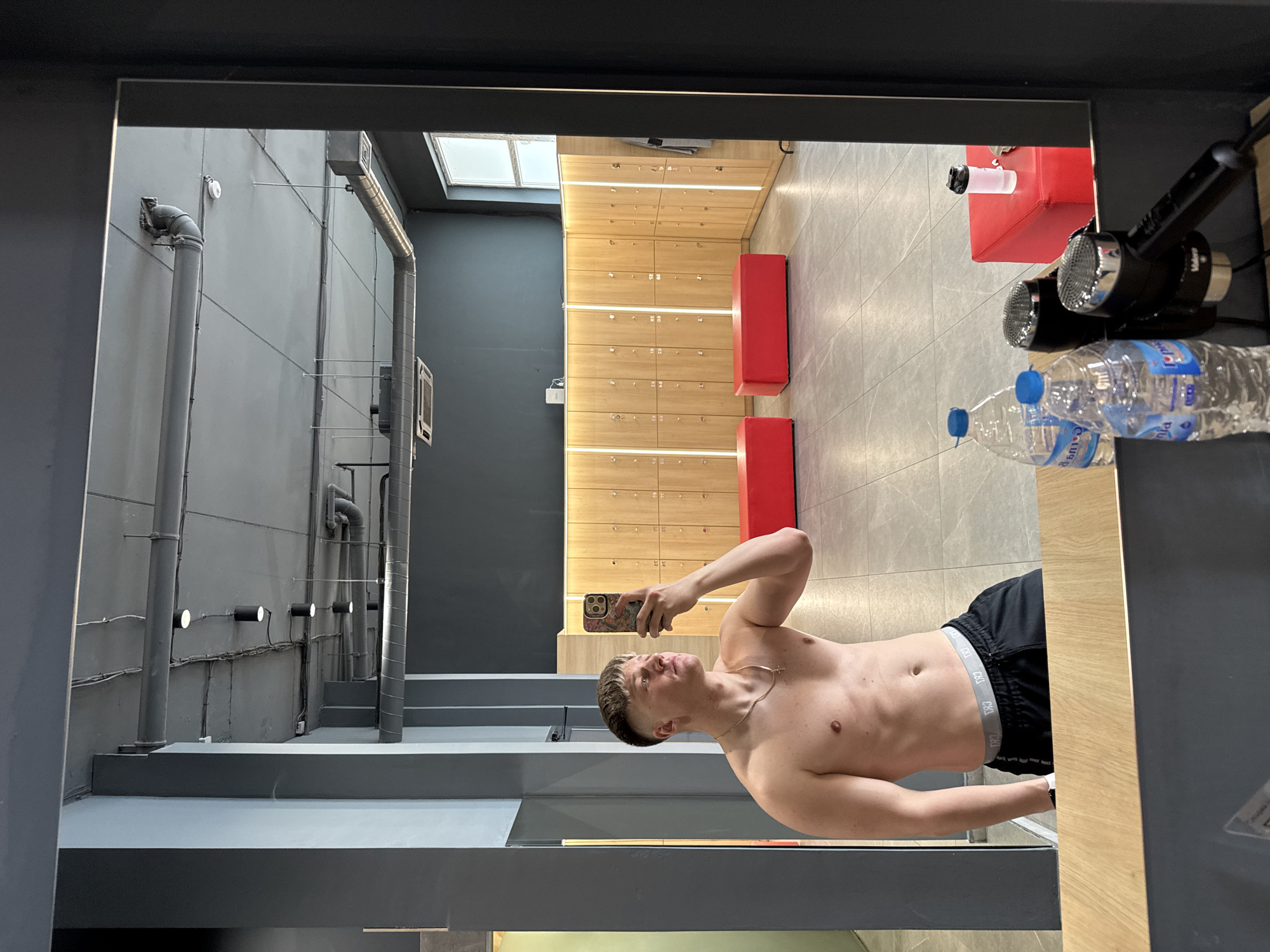 A person is taking a shirtless selfie in front of a mirror. The location appears to be a locker room with wooden lockers and red benches. The background shows gray walls with pipes and lighting fixtures. There are water bottles and hair dryers near the mirror. The photo may have been taken at a gym in Plovdiv, Bulgaria, but that cannot be determined definitively from the image alone.
