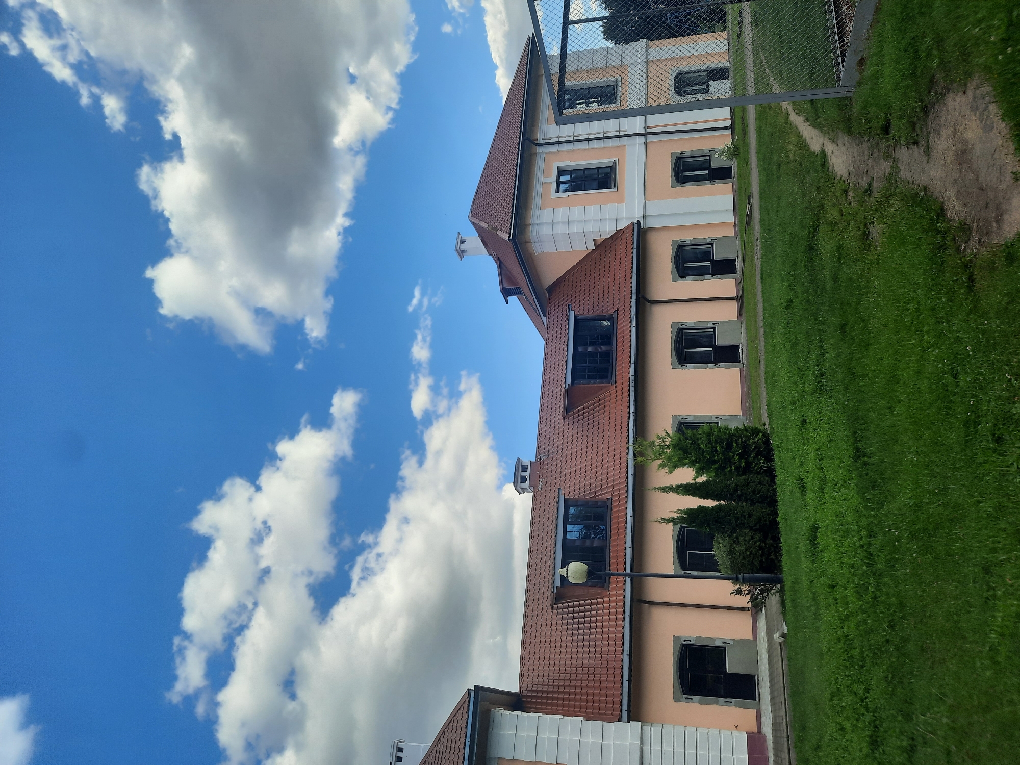 The image depicts the exterior of a pale orange building with a reddish-brown tiled roof. The building features white decorative trim around windows and corners, and several dark-framed windows are visible. Some of these windows are on the roofline, suggesting attic space. A metal fence runs along the right side of the image, bordering a grassy area with a dirt path. The sky is blue with scattered white clouds. The scene is likely located in Shklov, Belarus, given the provided context.