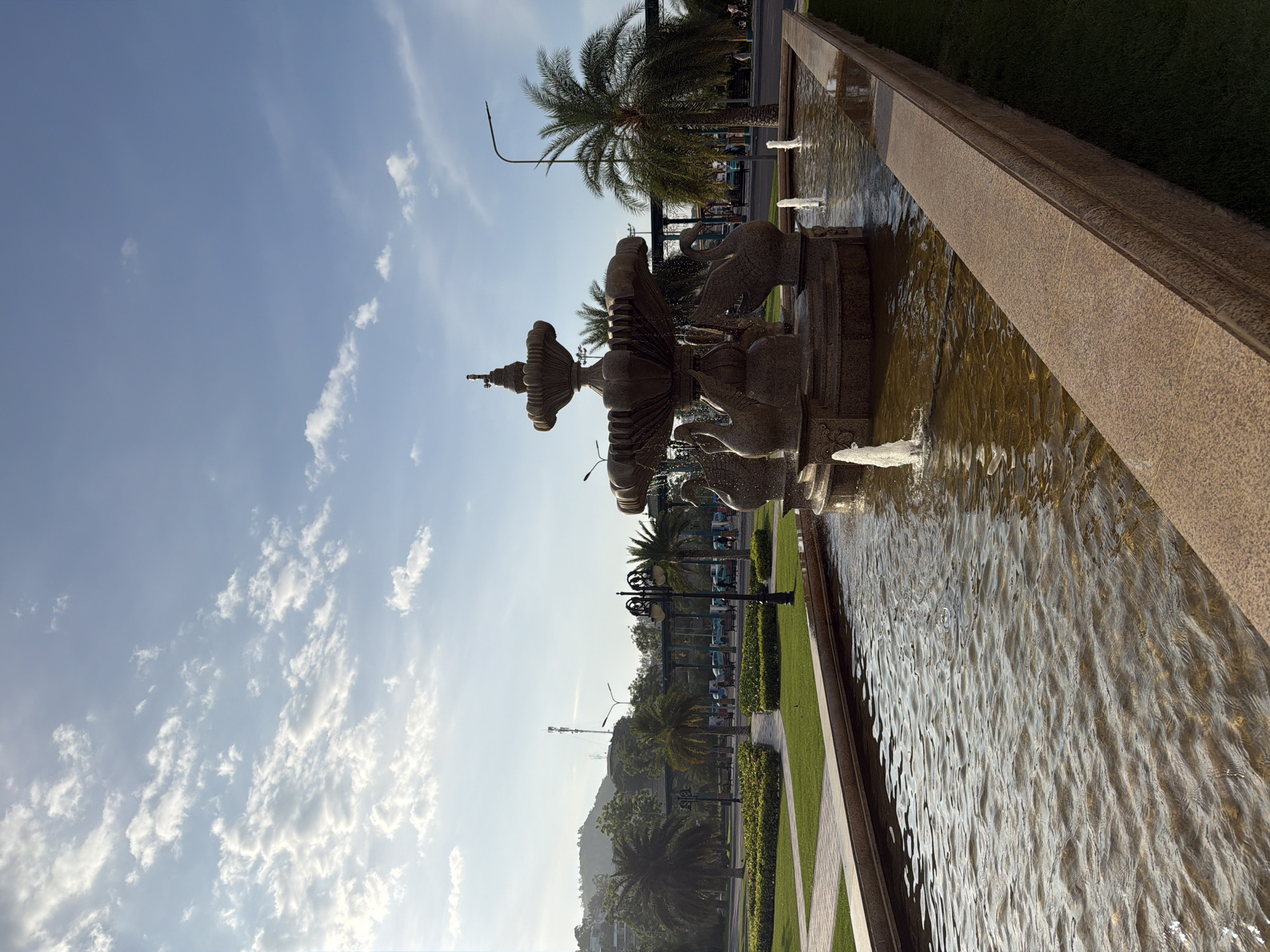 The image displays an outdoor public park or plaza located in Nha Trang Ward, Vietnam, under a clear blue sky with scattered white cumulus clouds, indicating bright daytime conditions. A prominent multi-tiered stone fountain, constructed from brownish-grey speckled granite-like material, occupies the middle ground. The fountain features sculpted swan figures around its lower tier and actively sprays water from its upper levels. This fountain is situated within a rectangular water canal, also made of the same stone, which extends towards the foreground and contains multiple smaller water jets producing vertical streams. The water in the canal is brownish-tinted and displays surface ripples.
To the left of the fountain and canal, manicured green lawns are segmented by paved pedestrian walkways. Several tall palm trees are interspersed throughout the park's landscaping. In the distant left background, a forested hill rises, topped by a slender communication tower. Multiple ornate black lampposts line the pathways. To the right, additional palm trees and modern streetlights on tall poles are visible. In the background, beyond the trees and a dark-columned covered walkway, several indistinct human figures can be seen, some appearing to interact near a series of light blue vehicles, possibly electric shuttles or golf cart-style taxis. The scene is illuminated by strong, warm sunlight from the upper left, creating distinct shadows.