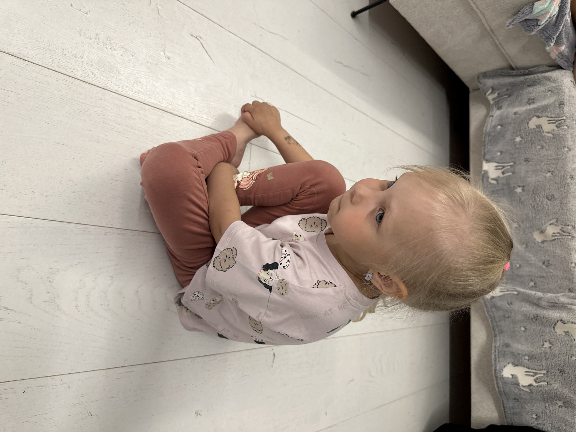 A young blonde-haired female child, estimated to be between two and four years old, is seated cross-legged on a light-colored wooden plank floor. She wears a light pink short-sleeved t-shirt patterned with various cartoon dog faces and muted reddish-brown leggings. A small pink hair tie secures a portion of her hair at the crown of her head, and she has small, light-colored stud earrings in her pierced ears. A faint temporary tattoo is visible on her right forearm, and a butterfly-like design is present on the upper thigh of her left legging. Her gaze is directed downwards and to the left, conveying a pensive or solemn expression. In the background, a light grey upholstered couch or bed is partially visible, covered by a grey fuzzy blanket featuring white, stylized patterns resembling hand gestures and stars. The scene is set indoors, likely in a residential dwelling in Прикамский, Russia.