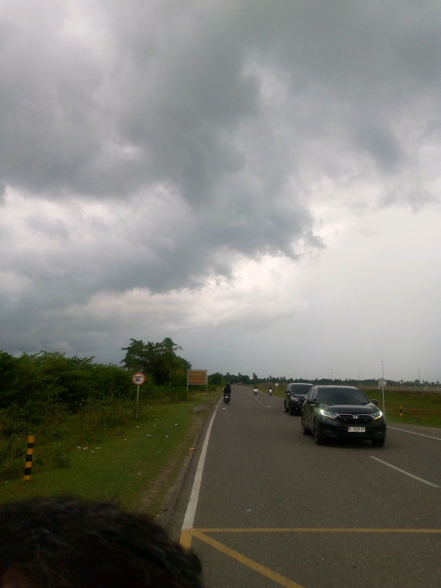 The image depicts a cloudy day scene on a paved road, likely in Glokaron, Indonesia, as suggested by the license plate of one of the vehicles. A dark SUV, identified by the license plate "BL 1674 ZF", is in the foreground, driving on the right side of the road. Behind it, another dark SUV and several motorcycles are also traveling in the same direction. The road is flanked by a grassy shoulder and dense green vegetation. A speed limit sign indicating "50 km/h" is visible on the left side of the road, along with a striped marker post. The sky is heavily overcast with dark, ominous clouds, suggesting impending rain. A sign is also visible on the right side of the road, but its text is unreadable. The overall atmosphere is one of a typical overcast day on a road in a semi-rural or suburban area.
