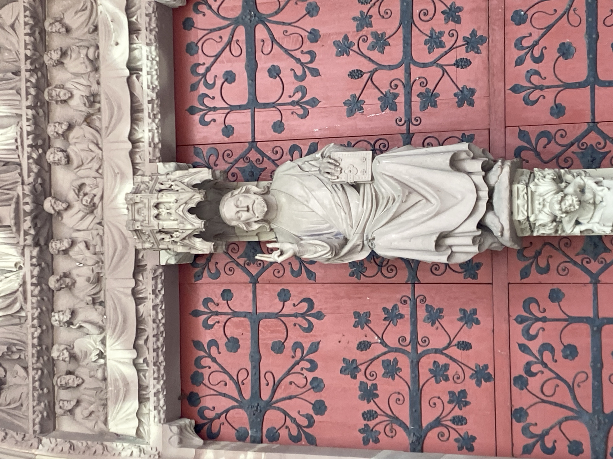 The image displays a close-up detail of religious statuary and architectural elements, likely from a cathedral in Strasbourg, France.
The central subject is a white stone statue of a male figure, depicted standing and holding an open book or tablet in his left hand, while his right hand is raised in a gesture, possibly of blessing or teaching. The figure has a beard and long hair, characteristic of depictions of Jesus Christ or a prominent saint, and is draped in flowing robes. He stands on an ornate pedestal, which merges into further sculptural details at the right edge of the frame. The figure's expression is serene and contemplative.
Behind the statue is a vibrant red wooden surface, which appears to be part of a large door or panel. This red background is extensively decorated with dark, almost black, wrought-iron style patterns. These patterns resemble intricate vines, leaves (some appearing like grape leaves), and stylized floral motifs or berries, creating a symmetrical and organic design across the panels.
To the left of the central statue, an elaborate architectural structure in a similar white stone is visible. This structure features several rows of smaller carved figures, primarily busts or heads, set within what appear to be niches or recesses of a column or pilaster. These figures suggest a larger sculptural ensemble, typical of a cathedral portal or facade.
The scene conveys a sense of historical artistry and religious grandeur. The lighting is even, suggesting either an indoor setting or a well-lit, perhaps overcast, outdoor environment, but no specific time of day or weather conditions are evident. No discernible text is legible on the book held by the statue or anywhere else in the image.
