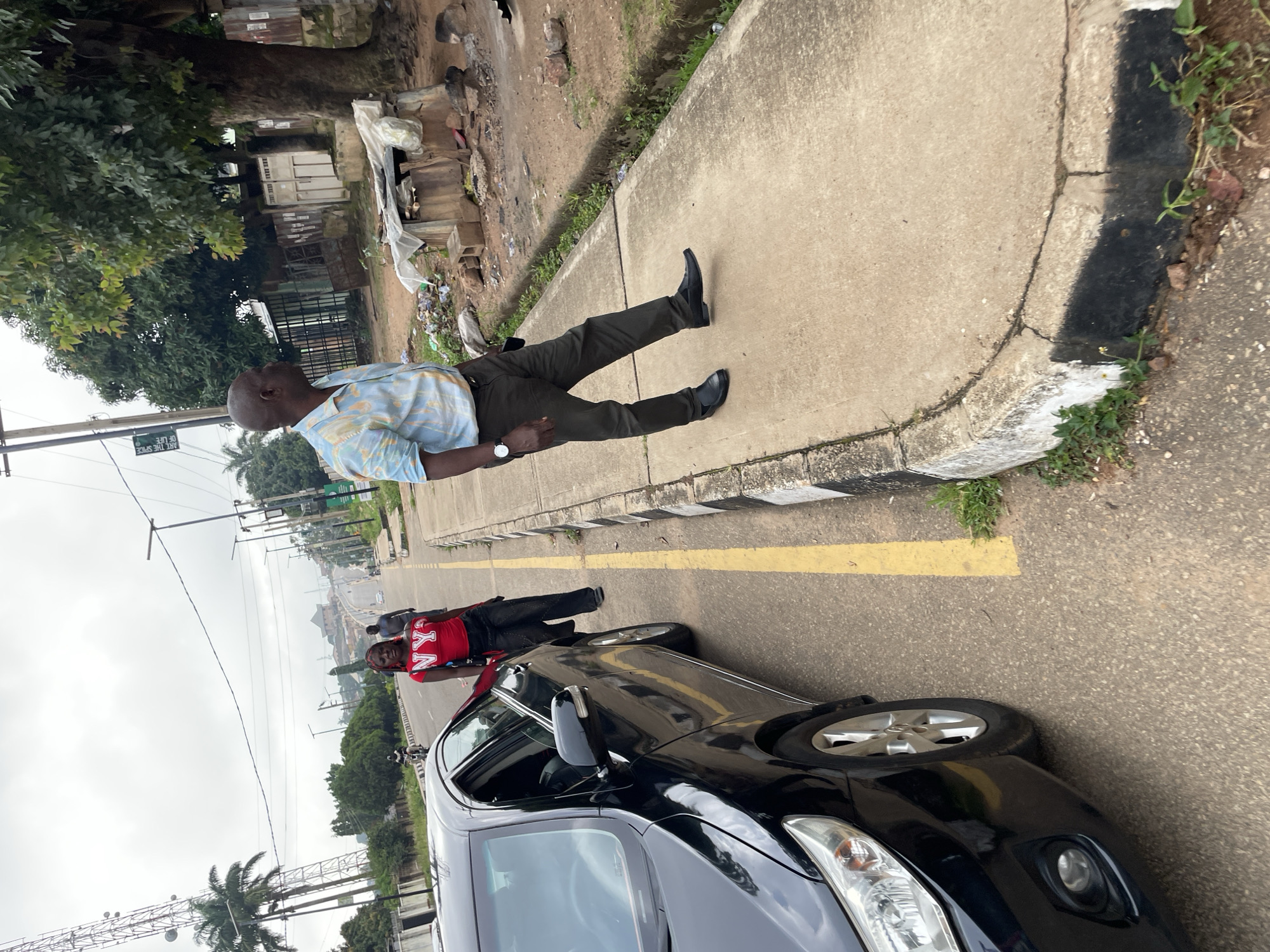 The image depicts a street scene in Jos, Nigeria, characterized by a cloudy sky and a paved road with a yellow line marking the lane. In the foreground, the left side of a black car is visible, with its door open. A young woman with red hair, wearing a red "NY" t-shirt and black pants, stands by the open car door. Further down the road, an adult male, dressed in a light blue and white patterned shirt and dark pants, walks away from the camera on the sidewalk. The sidewalk is bordered by a curb painted with black and white stripes. The background shows a street lined with trees and buildings, with some electrical poles and a large cell tower on the left. The environment appears to be urban, with signs of everyday life and infrastructure.