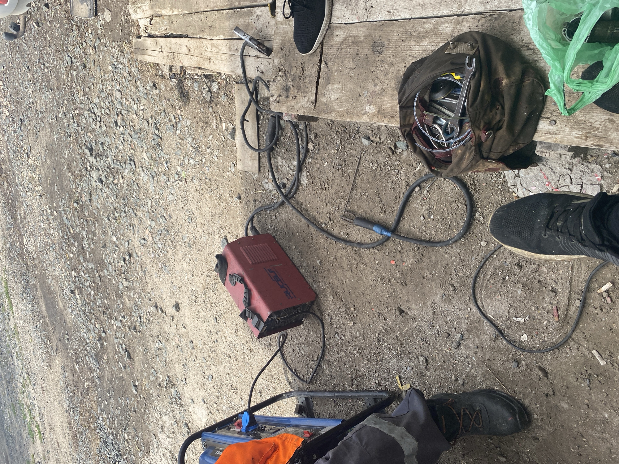 The image depicts an outdoor scene, possibly a workshop or construction site, with a gravel and dirt ground surface. In the foreground, a person's legs and feet are visible, wearing dark work boots. To the left, a blue generator with an orange safety vest draped over it is present. A red welding machine sits on the ground, connected by cables. To the right, wooden planks form a makeshift platform. A brown canvas tool bag filled with various tools, including wrenches and pliers, rests on the planks. A green plastic bag is also situated on the planks. The overall impression is of a work area with welding equipment and tools. The location is identified as Oktyabrsk, Russia.