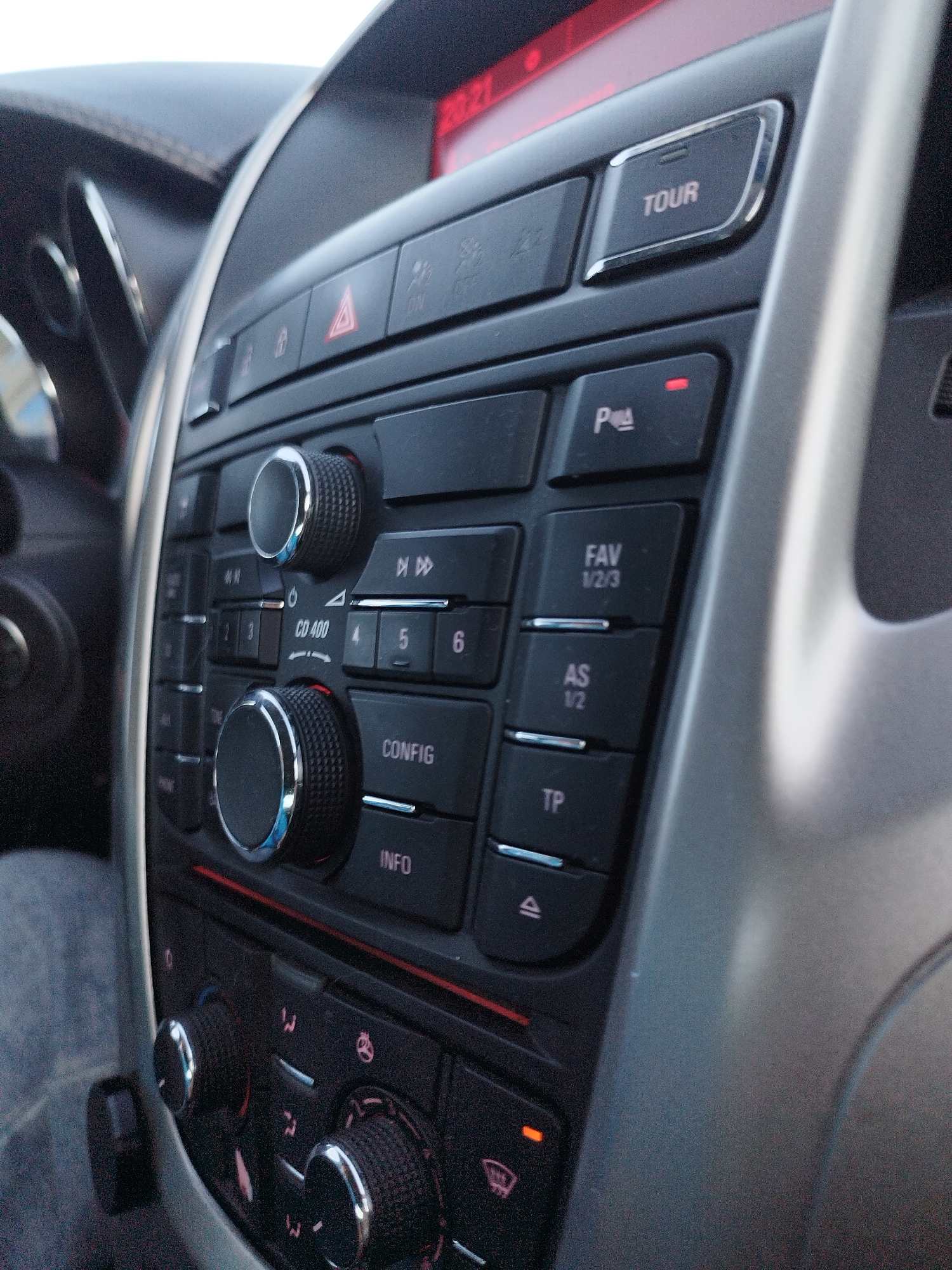 The image shows a close-up view of a car's center console, specifically the audio system and climate controls. The console is dark gray with some silver accents around buttons and knobs. Prominently visible are several buttons with labels like "TOUR," "P," "FAV 1/2/3," "AS 1/2," "TP," "CD 400," "CONFIG," and "INFO." There are also two large rotary knobs, one above the other, likely for volume and tuning. Below these controls are smaller knobs and buttons associated with the car's climate control system, indicated by symbols for fan speed, temperature, and air direction. A red LED light is visible on the "P" button. The overall environment is the interior of a car, with a portion of the dashboard and steering wheel visible in the background on the left, and a blurred silver trim piece on the right. The lighting suggests it is daytime, possibly with ambient light filtering into the car.