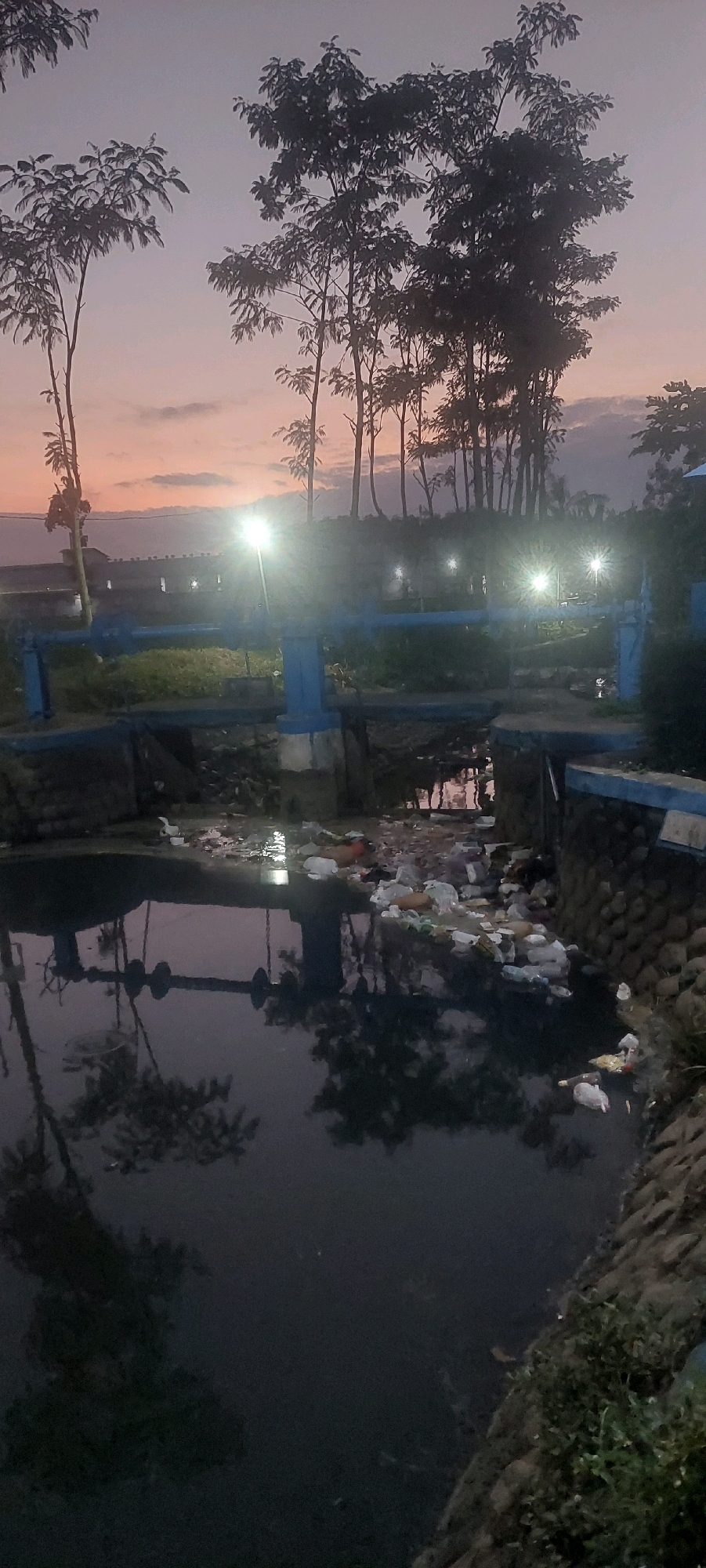 A scene in Jogoyudan, Indonesia at dusk shows a body of dark water filled with trash and debris, including plastic bags and bottles. The water is contained by stone walls and a concrete structure with blue railings. Trees with sparse foliage silhouetted against a pale orange and purple sky line the background. Several lights, likely streetlights, illuminate the area, reflecting in the water. A distant building with a few visible windows can be seen.