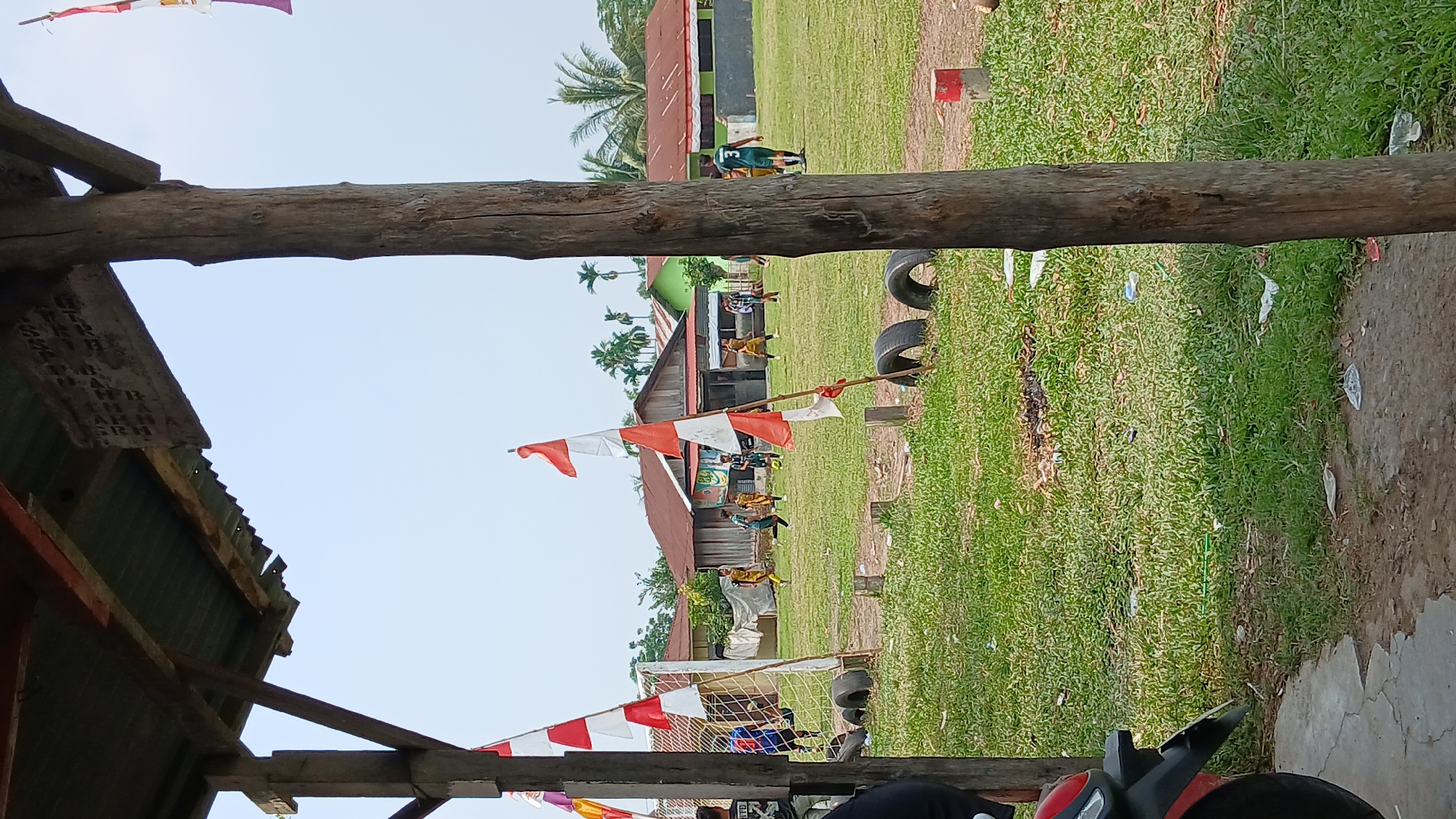 The image depicts an outdoor soccer field in an Indonesian village. The field is covered in green grass with some bare patches and scattered litter. Several Indonesian flags, red and white, are visible, some flying from poles near goalposts and others tied to makeshift barriers. In the background, traditional Indonesian houses with red or brown roofs are present. Several individuals are engaged in a soccer match, some in yellow and black uniforms, others in blue and black. A wooden structure with a corrugated metal roof is in the foreground, providing partial shelter. A motorcycle is partially visible on the left side of the image. The sky is clear and bright.