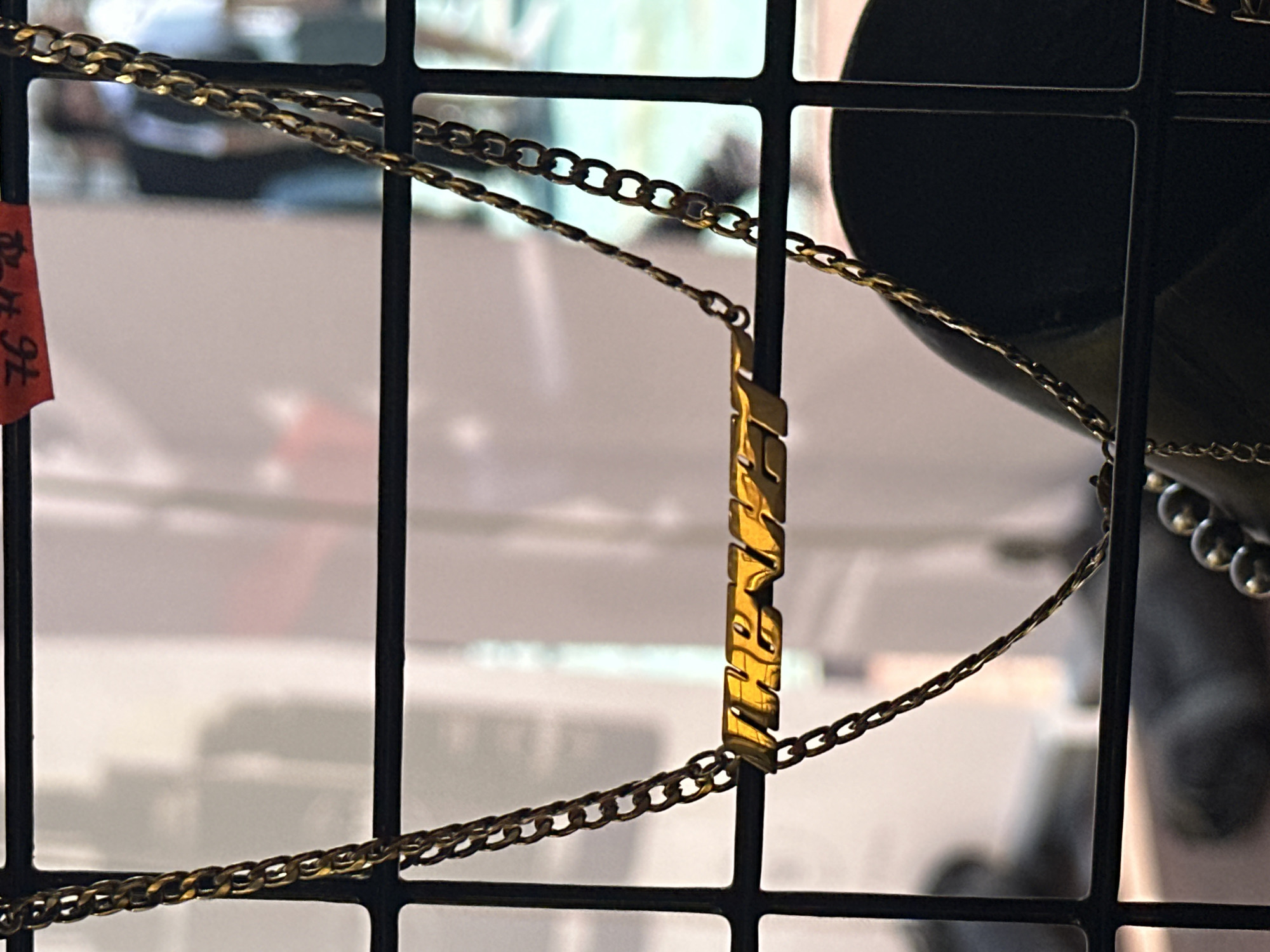 The image displays a gold-colored chain necklace with a horizontal bar pendant, suspended and viewed through a black metal grid, likely a display rack, in a retail setting in Sofia, Bulgaria. The pendant prominently features the word "THE WEEKND" in a stylized, reflective gold-toned font. In the upper right corner, a red price or identification tag is affixed to the grid, displaying the number "76" and partially visible Cyrillic characters. The background is blurred, showing indistinct figures of people, one appearing to wear a dark suit jacket and a lighter shirt. The lower right portion of the background reveals a dark, rounded object, potentially a jewelry display bust, with a strand of dark, spherical beads or pearls. The scene is brightly illuminated, creating highlights and reflections on the metallic components of the necklace.