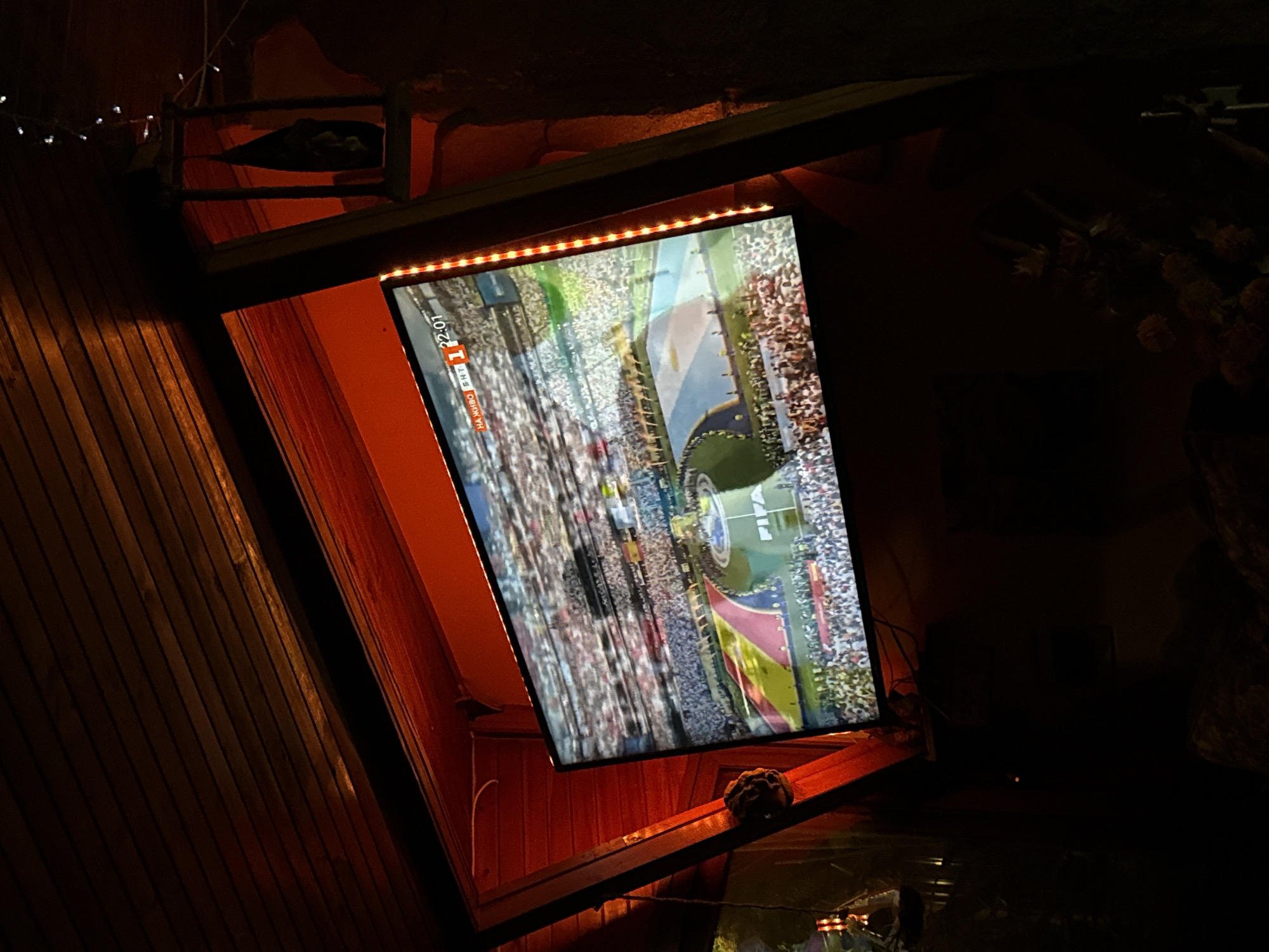 The image shows a television screen displaying a soccer match in a dimly lit indoor space with wooden paneling on the ceiling. The screen, positioned at an angle, shows a stadium filled with spectators and a soccer field with flags. A clock on the screen indicates "22:01" and text that appears to read "НА ЖИВО БНТ 1". Red LED strip lights are visible along the right side of the screen and framing the red-painted structure behind it. The overall impression is that of a bar or a covered outdoor seating area, possibly in Mineralni Bani, Bulgaria, designed for watching sports.