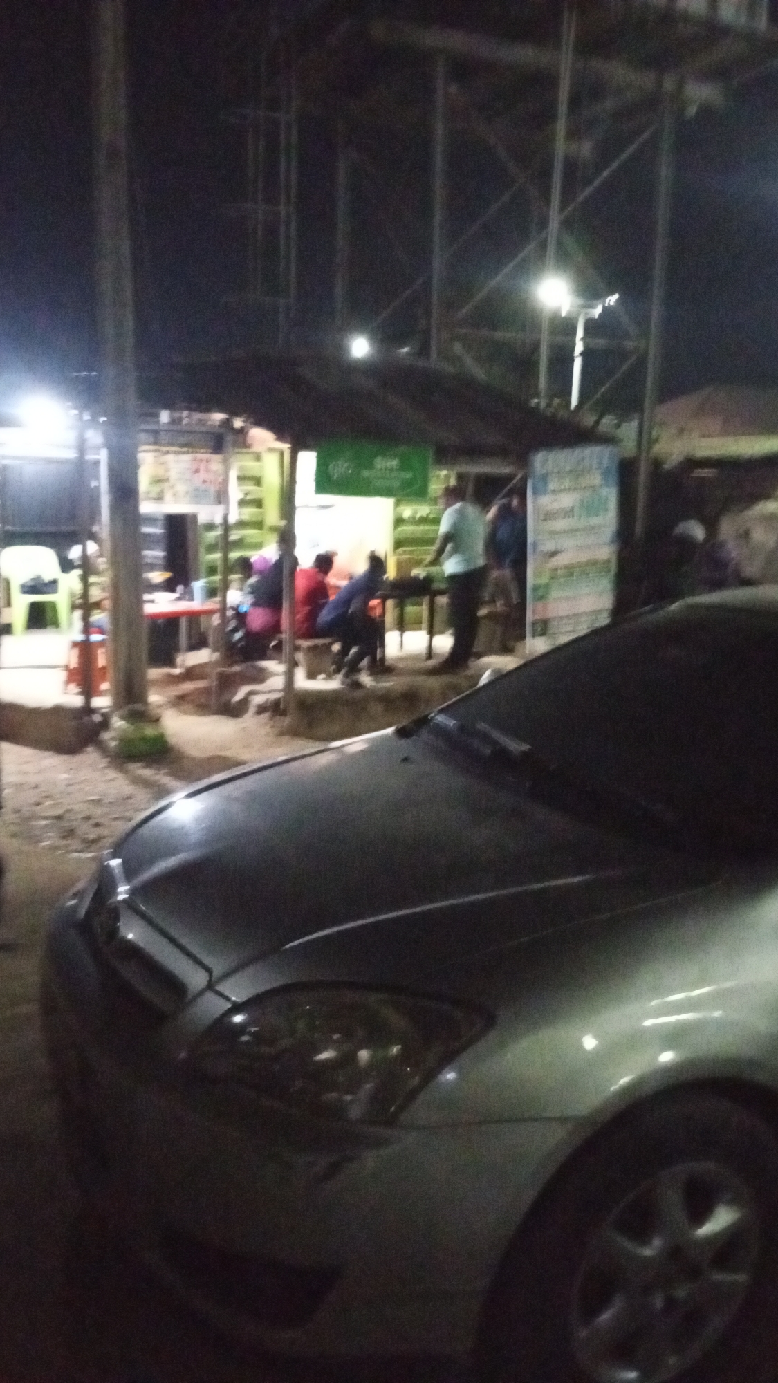 The image depicts a nighttime scene in Adankolo, Nigeria. A silver-colored car is parked in the foreground. Behind the car is a small, open-air eatery with a corrugated metal roof and a green sign. Several people are seated at tables, some are standing. Illumination comes from overhead lights and the headlights of the car. A utility pole is visible on the left side of the frame. Steel communication towers are in the background.