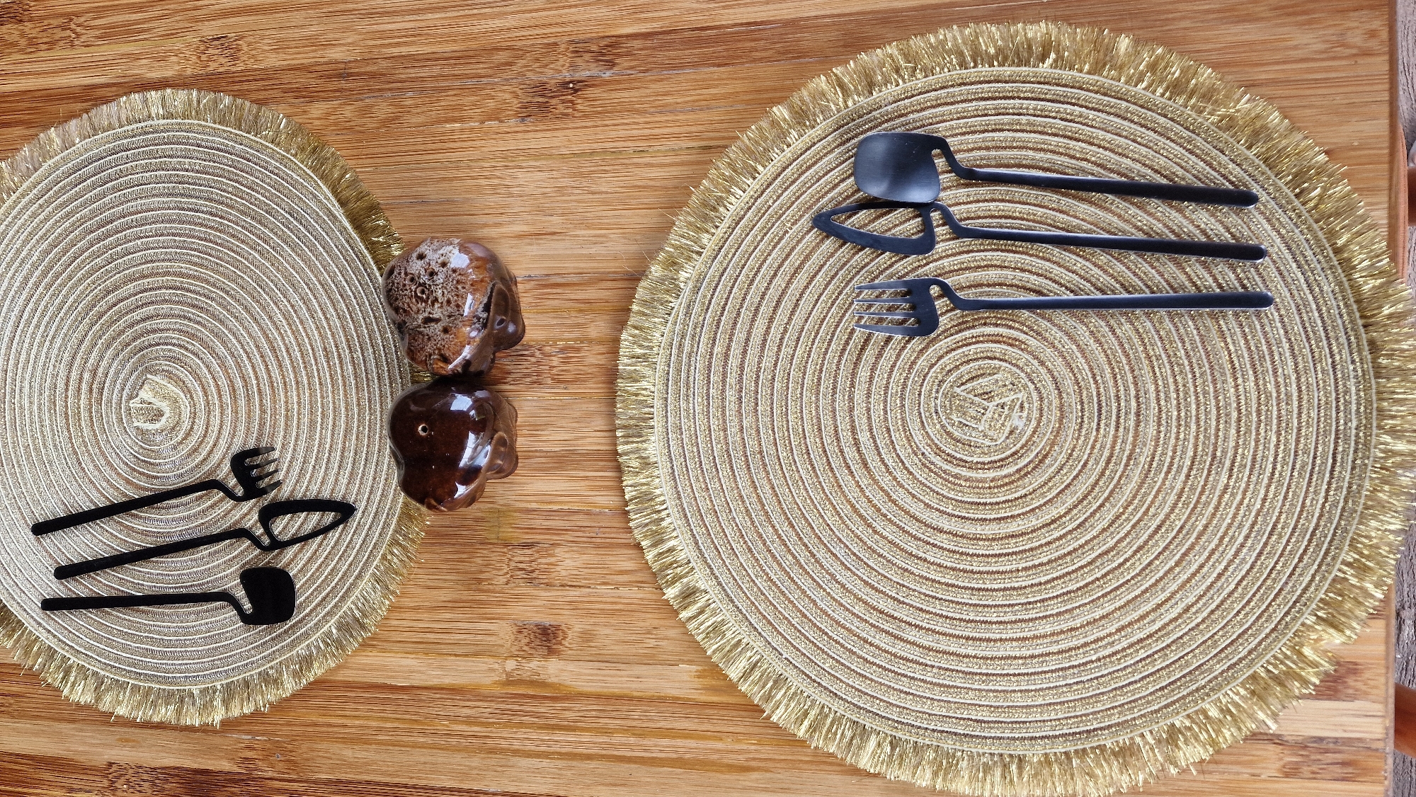 The image displays a meticulously arranged table setting on a light-colored wooden surface, likely bamboo, within the city of Baan Mae Nam, Thailand. Two round, woven placemats, shimmering gold with concentric patterns and a fringed edge, are present.
On the upper placemat, three black, novelty utensils are arranged: a small, flat-bladed shovel, a pointed spade, and a three-tined rake, designed to resemble miniature gardening tools.
Between the placemats, on the wooden table, sit two small, polished, ceramic or stone decorative objects. The left object is a dark brown, smooth, bulbous form, while the right object is a similar shape with a mottled brown and cream, textured surface.
On the lower placemat, three black, modern-design dining utensils are positioned: a four-tined fork, a distinctive blade-shaped spreader, and a wide-bowled spoon. No people or human activity are visible in the scene.