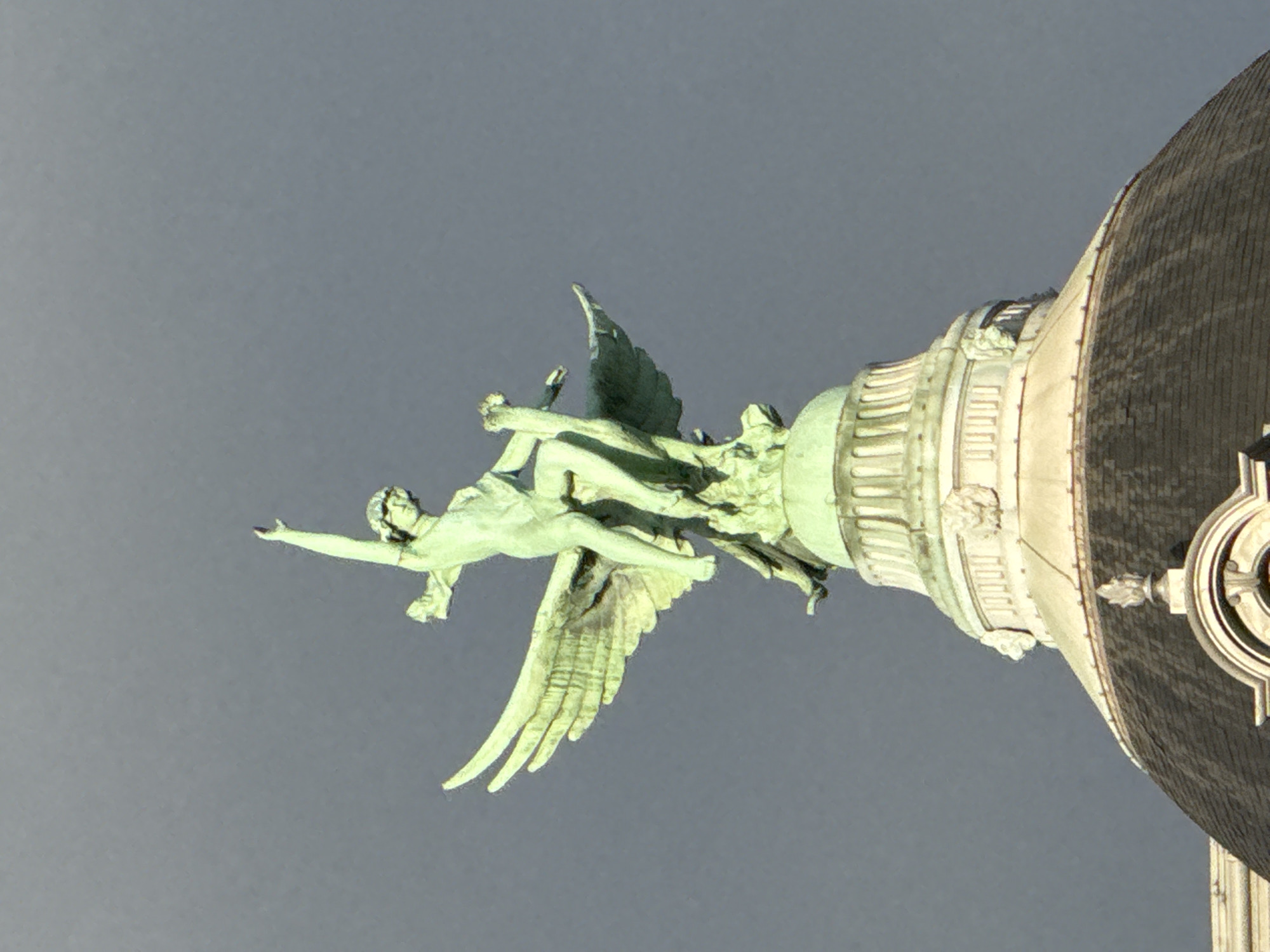 The image displays a patinated green statue of an angelic or mythological figure against a plain, overcast grey sky. The sculpture is an ornamental part of a large, classical architectural structure, likely a dome or cupola of a grand building.
The main subject is a winged, male-like figure, possibly an angel or a classical deity, crafted from aged bronze or copper, evident from its distinct greenish patina. The figure is captured in a dynamic, outstretched pose, as if in mid-flight or launching forward, with arms extended and legs bent. Its wings are spread behind it. A second, less prominent figure is visible behind the primary one, seemingly part of the same sculptural ensemble, possibly supporting or interacting with the main figure.
The statue is affixed to an ornate, multi-tiered architectural element that forms the base of a larger, darker dome. This base is richly detailed with cornices, decorative friezes, and what appear to be sculpted heads or small figures. The dome itself is dark, possibly covered in slate or metal, and has a curved profile.
The sculpture vividly portrays a moment of dramatic motion, suggesting an allegorical or mythological narrative. The uniform grey sky indicates an overcast day, which serves as a neutral backdrop, accentuating the intricate details and the vibrant green hue of the statue. The perspective is from below, looking upwards, highlighting the grandeur and scale of both the sculpture and the architecture. No text is visible in the image. Given the provided context of Paris, France, the architectural style and monumental scale are characteristic of significant landmarks such as the Opéra Garnier or other prominent 19th-century buildings in the city.