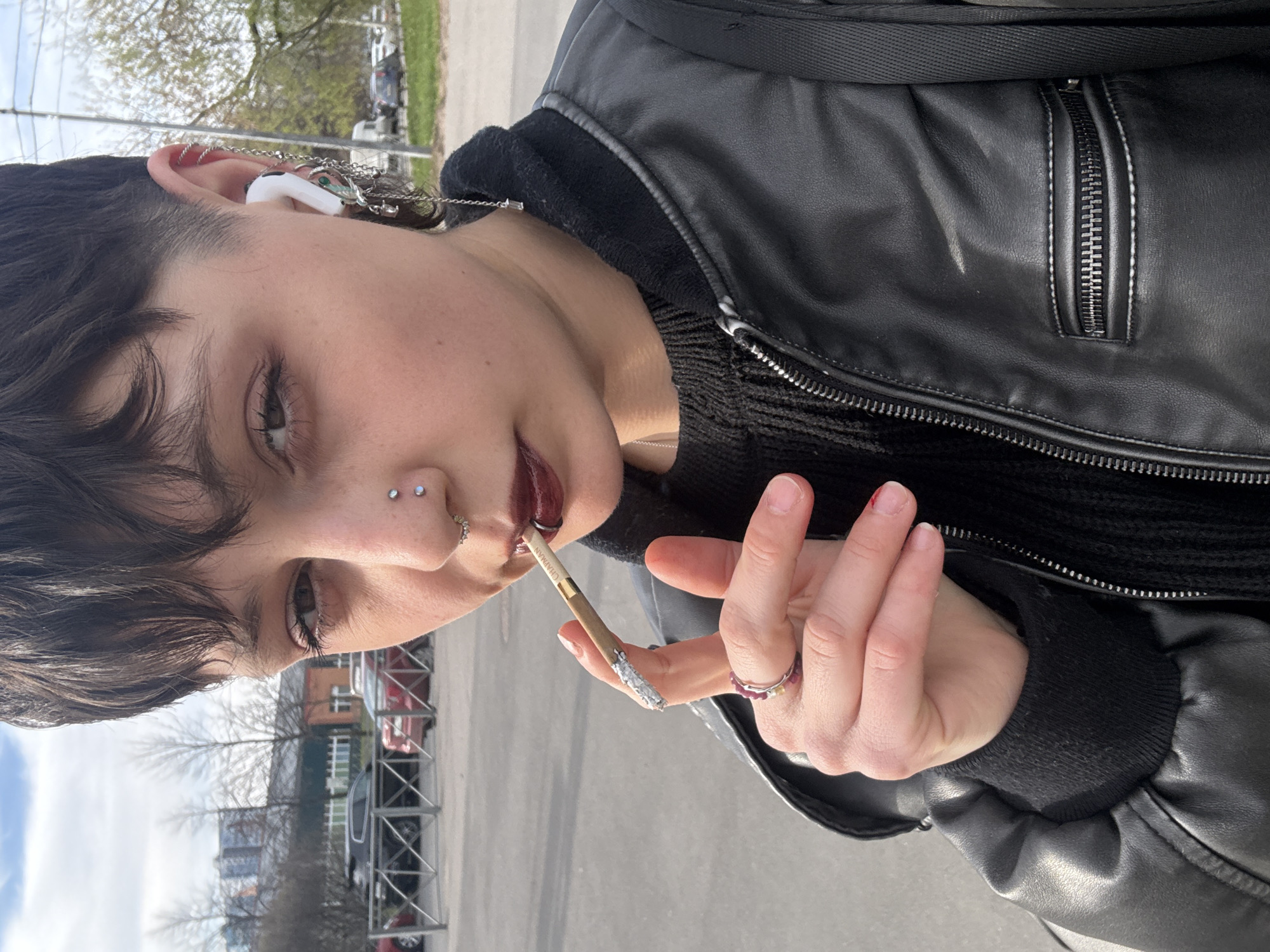 A person with short brown hair, fair skin, and multiple facial piercings is holding a lit cigarette, possibly a clove cigarette, in their right hand. They are wearing a black leather jacket, a black knitted sweater, and a silver ring on their right index finger and another ring on their right pinky finger. The person is also wearing a white wireless earbud in their left ear, which is adorned with a chain. The background shows a city street with parked cars, bare trees, and buildings. It is daytime, likely in the spring, as indicated by the budding trees.
The image was not taken in the city of Saint Petersburg, Russia.