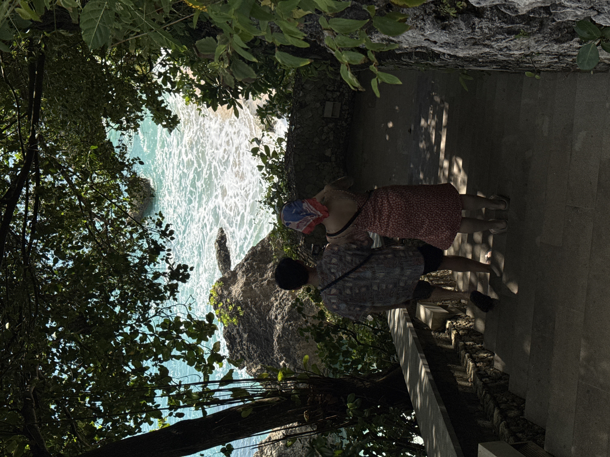 Captured from an elevated perspective, the image showcases a coastal scene in Kuta Selatan, Indonesia. The composition is framed by verdant foliage in the foreground, partially obscuring a turquoise sea with white-crested waves crashing against a rocky outcrop. Two individuals, one wearing a reddish patterned dress and a face covering, and the other in a patterned shirt, are standing on a paved pathway adjacent to the scene. The pathway is bordered by a stone wall, casting shadows onto the steps. The environment is characterized by a blend of natural and man-made elements.