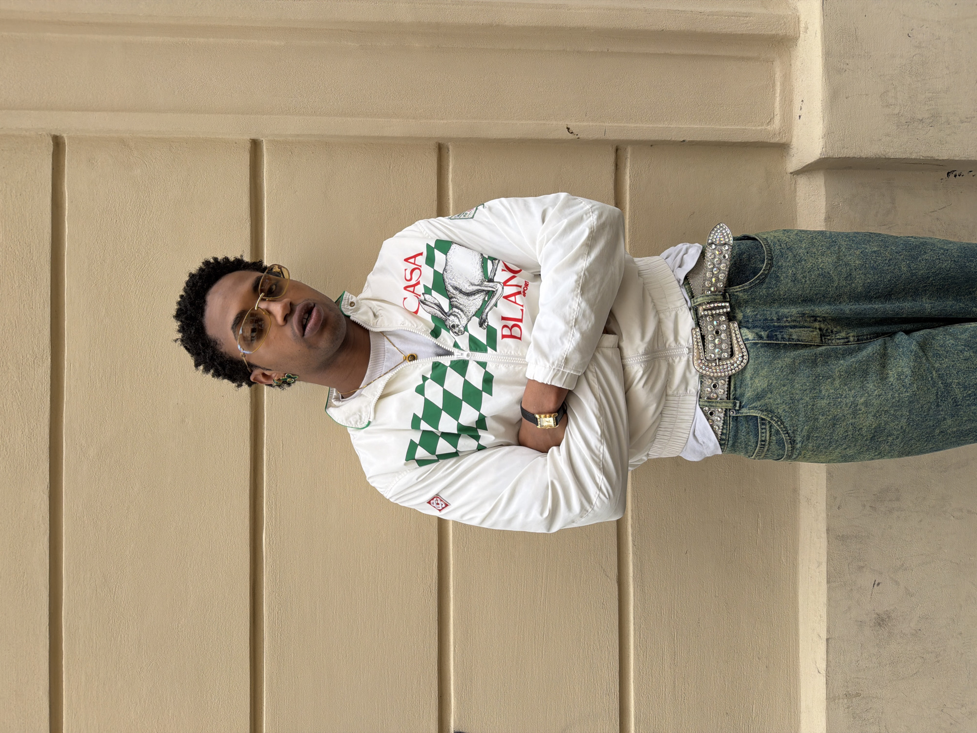 A young man poses confidently against a textured, light beige wall, characteristic of Parisian architecture. He stands with his head slightly tilted and his hands tucked into his pockets, looking directly towards the viewer through stylish yellow-tinted sunglasses. He has short, dark, curly hair.
He is wearing a white track jacket adorned with a prominent green and white checkered pattern across the chest and back. On the front, in red text, are the words "CASA BLANCA SPORT" accompanied by a graphic of a white rabbit. Beneath the jacket, a simple gold-toned chain is visible. On his left wrist, he sports a gold-toned square watch with a black leather strap. He pairs the jacket with acid-wash blue jeans and a distinctive wide, sparkling silver belt featuring an ornate buckle, all heavily embellished with rhinestones.
The lighting suggests it is daytime, and the weather appears clear. The wall behind him has subtle vertical indentations, adding to the architectural detail of the setting. The man's expression, partially obscured by his sunglasses, conveys a sense of cool confidence.