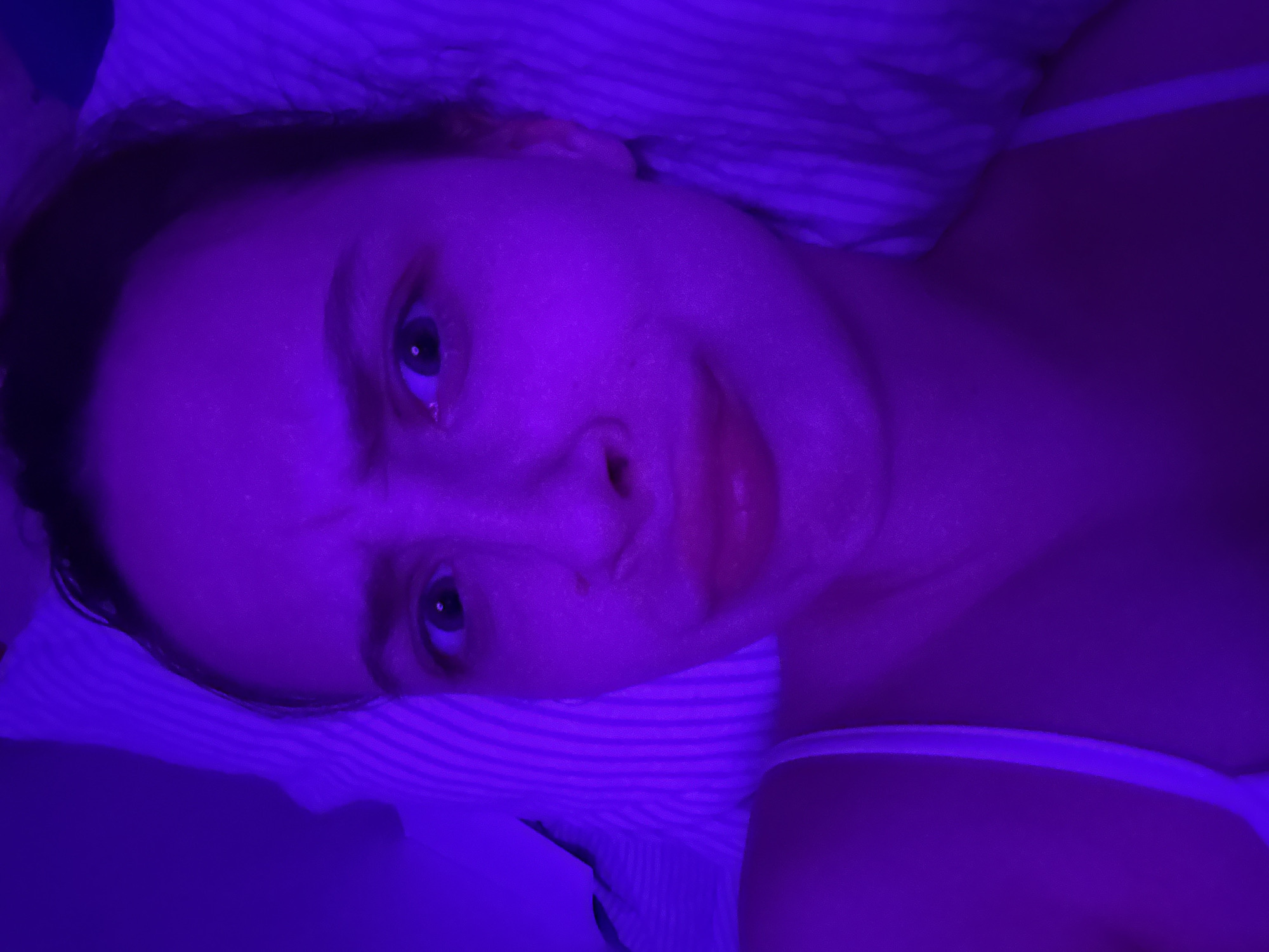 A woman is lying down in a bed with striped bedding. She is wearing a white tank top. The image is illuminated with blue light. It is impossible to determine the location of Прикамский, Russia, from this image.