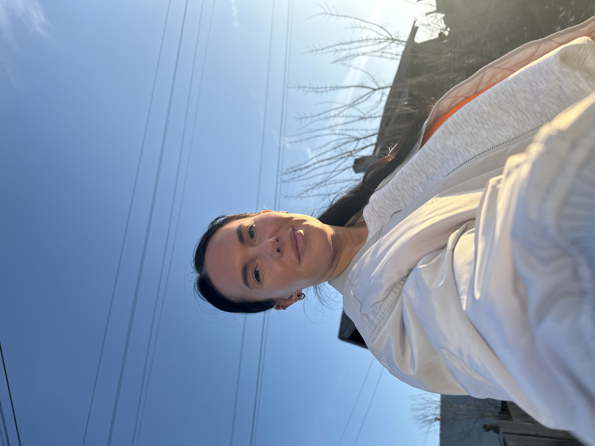 A woman is captured in a close-up outdoor portrait, looking directly at the camera. She has dark hair, styled away from her face, and a neutral to slightly pleasant expression. She appears to be wearing a light-colored, possibly off-white or light grey, jacket over a grey garment, with a hint of an orange layer underneath. Small, dark earrings are visible on her ears.
The setting is outdoors, dominated by a clear, bright blue sky in the background. Several dark, thin power lines stretch across the sky, running horizontally across the middle and lower portion of the frame if the image were properly oriented. In the upper right quadrant of the background, intricate bare tree branches are visible against the sky, suggesting winter or early spring. A darker structure, possibly part of a building or fence, is partially obscured behind the branches.
The scene is brightly lit by natural sunlight, creating a slight glare or haze in the upper right, indicating the sun's position. This illumination also creates highlights on the woman's clothing. The image appears to be a selfie or a close-up shot taken by someone else, capturing a moment under a clear sky in what appears to be a rural or suburban environment in Olkhovka, Russia. There is no visible text in the image.
