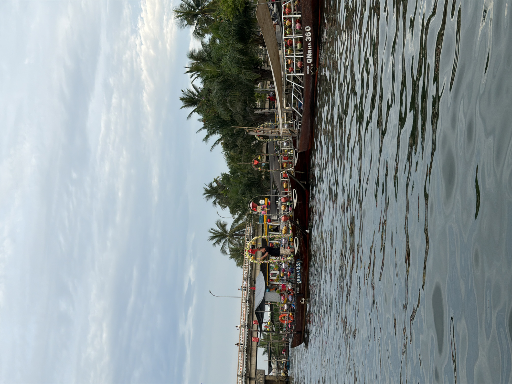 This image depicts a scenic view of a river or canal in Hội An, Vietnam, under an overcast sky, likely during late afternoon or early evening. The foreground and lower portion of the image are dominated by the dark, rippling water of the waterway, which reflects the soft, diffused light from the sky.
The central subjects are several traditional wooden boats moored along the riverbank. These boats are richly adorned with numerous colorful lanterns in shades of red, yellow, green, and blue, as well as festive garlands and decorations, suggesting a celebratory occasion or tourist activity. One boat in the mid-ground clearly shows a person standing on its deck, possibly a crew member adjusting decorations. Other smaller figures can be seen on different boats and on the bridge in the background.
Behind the boats, the riverbank is covered in dense, lush tropical foliage, including prominent tall palm trees. A concrete bridge with white railings and lampposts spans the waterway in the distance.
On the side of one of the decorated boats, the text "QNA HA 360" is clearly visible, with "GOUCH VOB" written beneath it. Another boat features partially visible text "QNAHA32", likely indicating similar registration details. The festive decorations, traditional boats, and tropical setting are strong indicators of Hội An's unique charm, particularly its renowned lantern culture and river cruises. The overall atmosphere appears calm and enchanting.