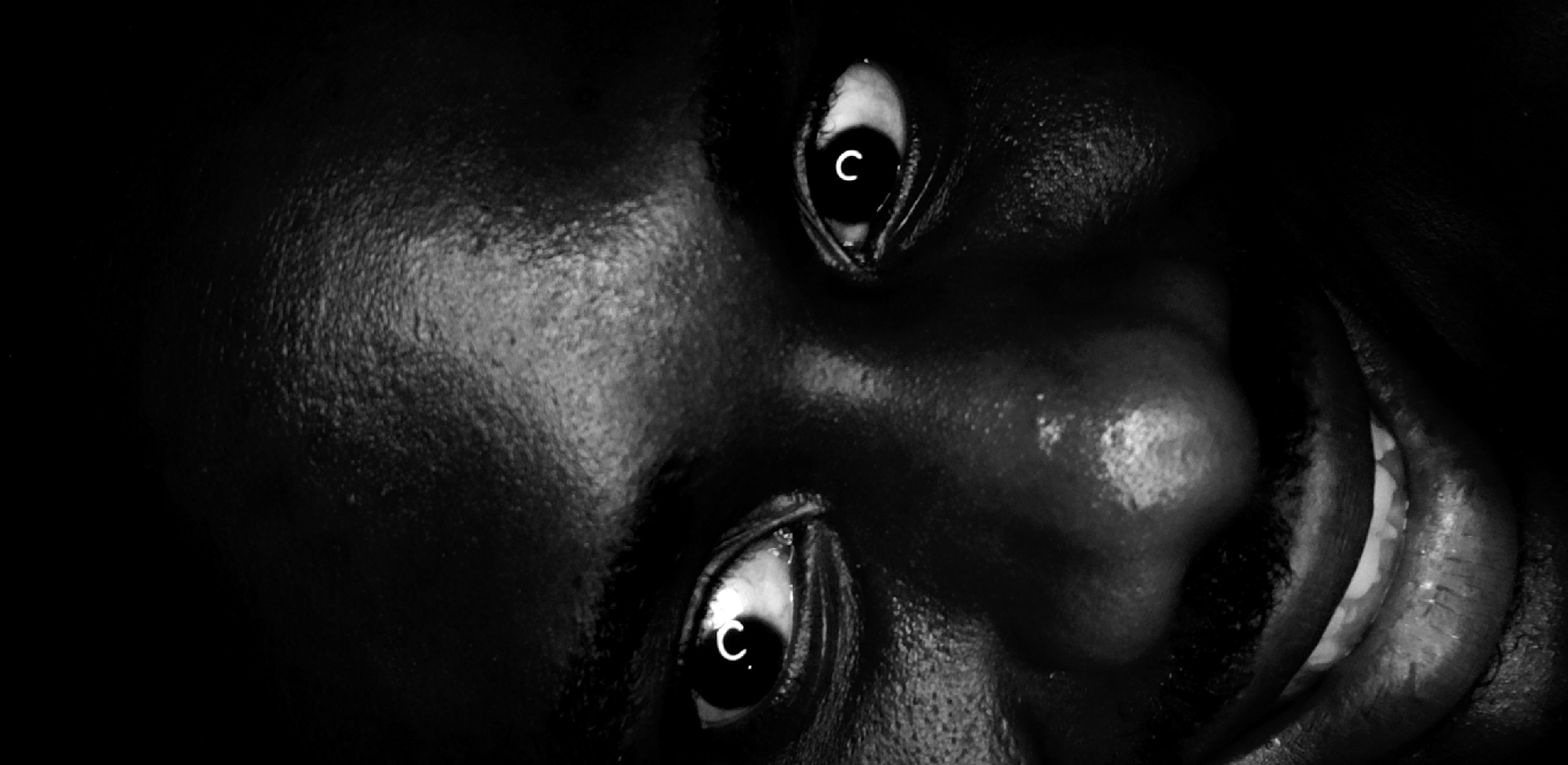 A high-contrast, black and white close-up image shows the face of a person, likely male, with dark skin, oriented diagonally across the frame. The subject's head is tilted, presenting the face from an upward angle, almost as if looking down at the viewer or from a supine position.
The most prominent features are the wide-open eyes, both with bright white sclera and dark irises/pupils. In the center of each pupil, a distinct white letter "C" is visible, likely a reflection from a light source or an object in the environment. The skin, particularly around the cheeks, forehead, nose, and lips, shows a reflective, possibly moist or oily texture, highlighting the contours of the face under dramatic lighting.
The nose and a portion of the mouth are visible towards the lower right of the frame. The mouth is slightly open, revealing bright white teeth in what appears to be a subtle or intense smile, or perhaps a grin. Deep shadows consume much of the background and parts of the face, emphasizing the stark contrasts and creating a dramatic, somewhat unsettling mood. The overall expression suggests intensity or focused attention, with a hint of an enigmatic emotion. No explicit location cues are visible within the image, but the subject's features are consistent with a person of African descent, aligning with the context of Narayi, Nigeria.