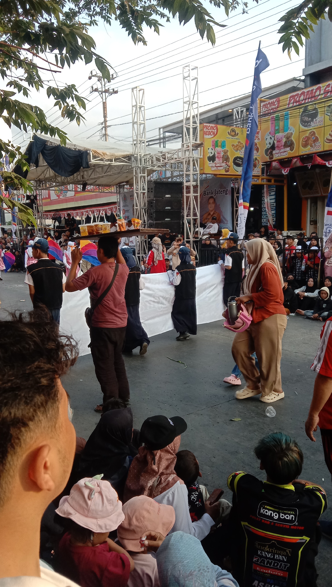 In Jatisawit, Indonesia, a street scene unfolds with a crowd gathered for an event, possibly a festival or market, as indicated by the presence of a stage setup with speakers and scaffolding. A man in a maroon t-shirt and dark pants carries a tray of drinks balanced on his head, walking through the throng. Several individuals, some wearing headscarves, are visible, including a woman in an orange top and tan pants, and others in black clothing. In the background, banners promoting "PROMO HEBOH" and "Bank Jateng" suggest commercial activity. The ground is paved, and the event appears to be taking place outdoors, with natural light filtering through the leaves of trees overhead. A group of people, including children, are seated on the ground, some observing the activity. The atmosphere seems festive and communal, with a mix of participants and spectators.