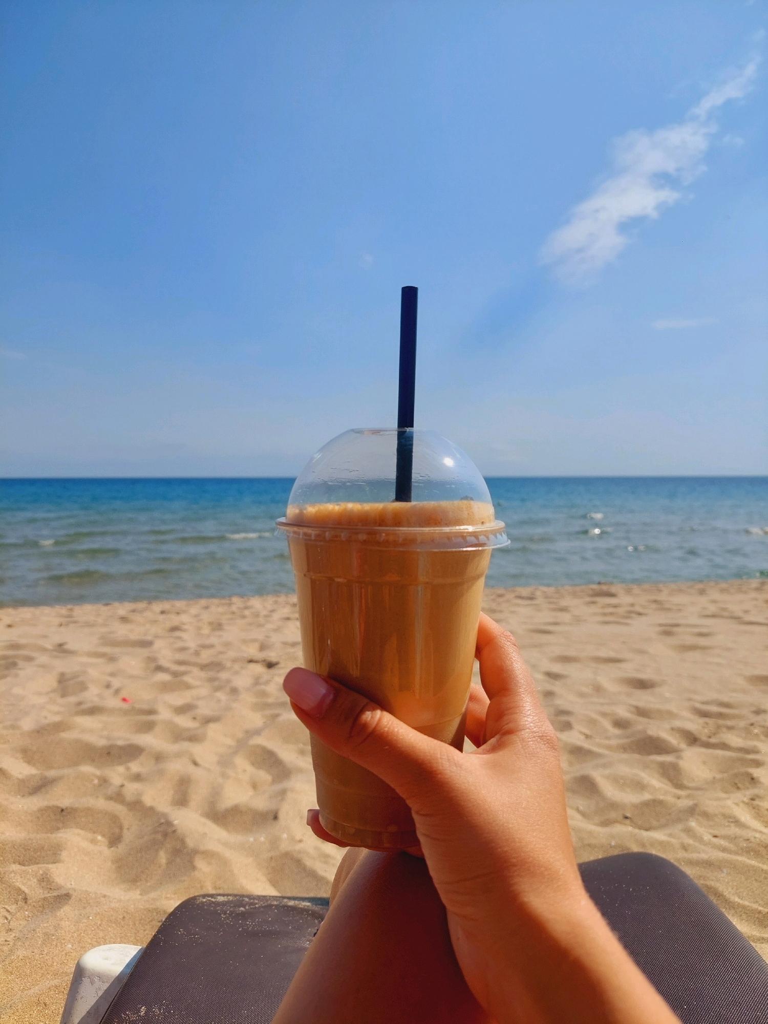 A close-up, eye-level shot shows a person's hand holding a clear plastic cup filled with a light brown beverage, likely coffee or a milkshake, with a black straw. The cup has a clear plastic lid. The hand has manicured fingernails painted pink. The background is out of focus, showing a sandy beach and the blue sea with a clear blue sky above. The scene appears to be set in the city of Sozopol, Bulgaria.