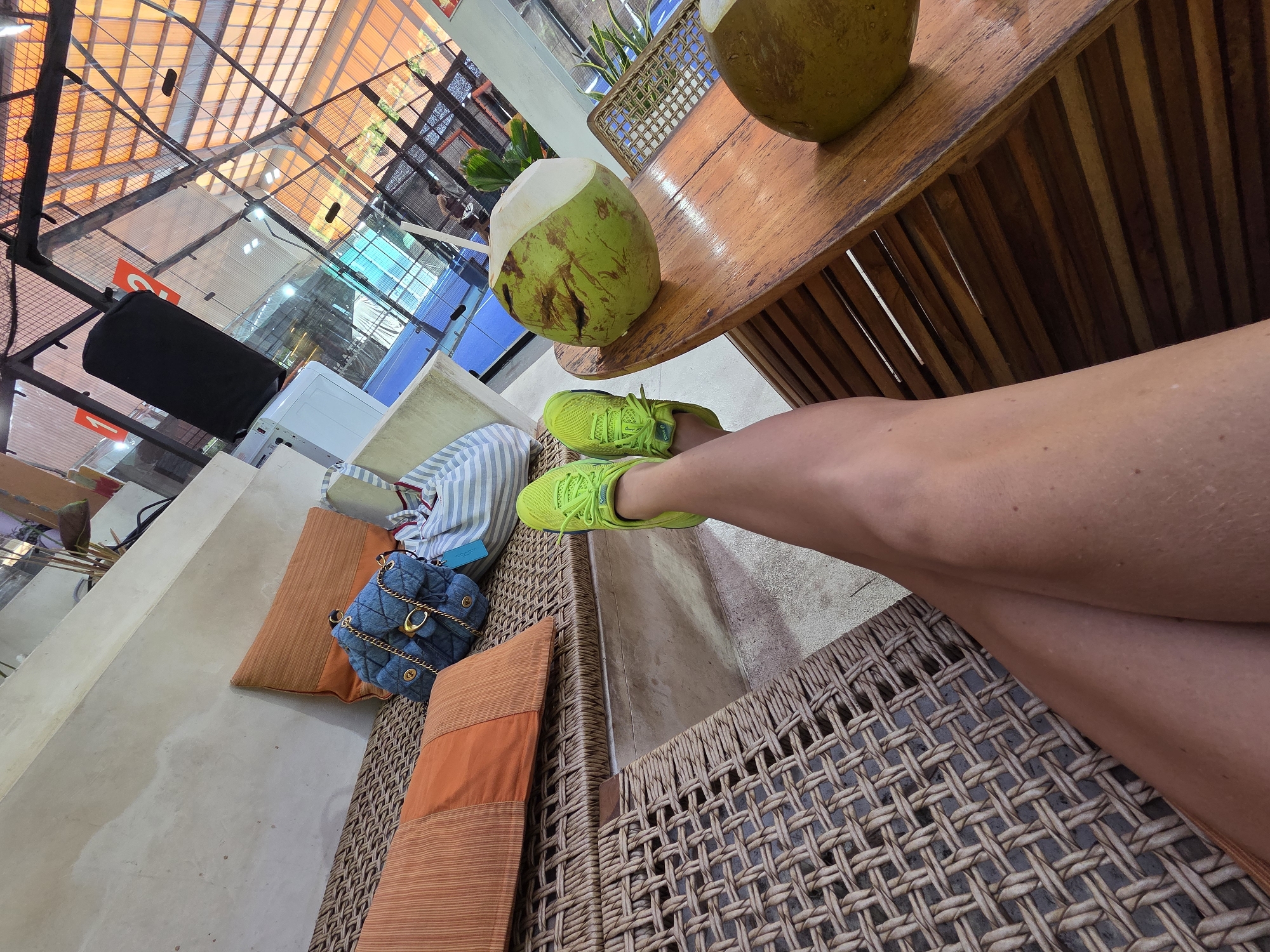 A person's legs, clad in bright green sneakers, are crossed and resting on a woven wicker lounger in a covered outdoor seating area. The person's legs dominate the foreground, with one leg extending towards the camera. Behind the lounger, a blue quilted handbag with gold hardware and a striped tote bag are visible. Orange cushions are arranged on the lounger. In the background, a wooden table holds a halved green coconut with a straw. Beyond the seating area, a blue sports court is visible through a glass partition, with a sloped orange roof structure overhead. The location is the lobby area of a sports facility in North Kuta, Indonesia.