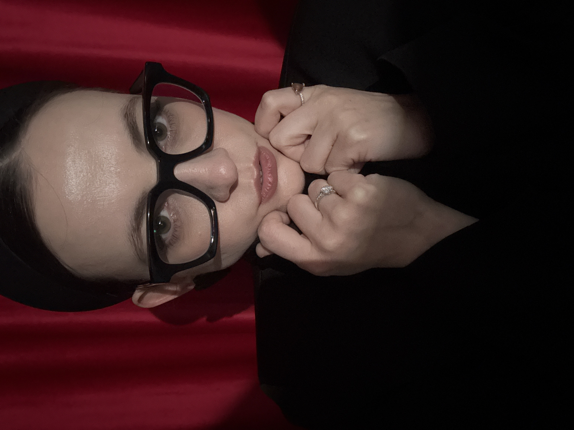 A person with fair skin, wearing black-framed glasses, is photographed from a close, angled perspective against a red and black backdrop. The person's fingers are pressing into their cheeks, revealing two rings on their hands. The environment is dimly lit, focusing attention on the subject's face. The image does not provide any information about the location being the city of Chaikovsky, Russia.