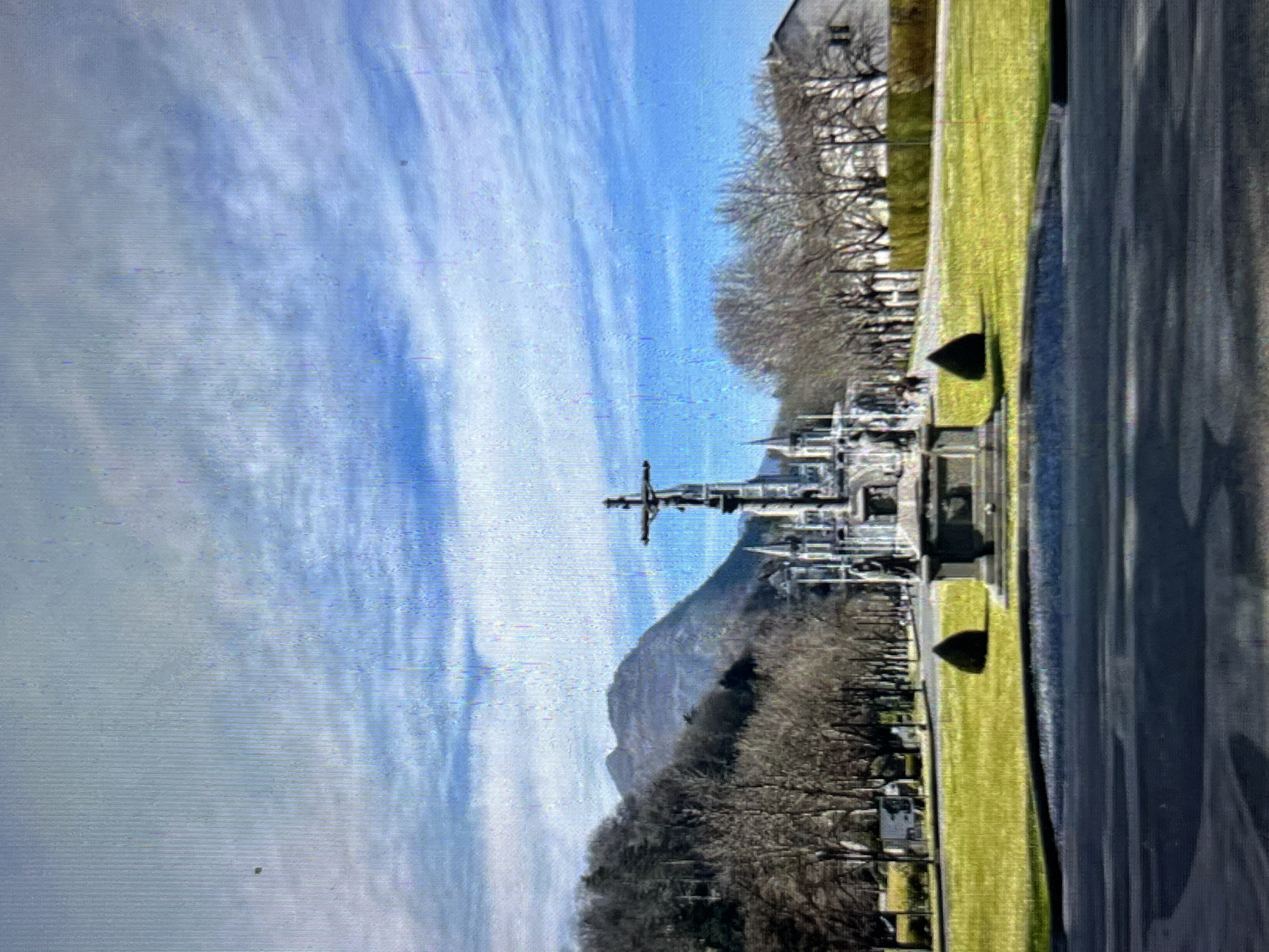 The image shows a landscape in Tinaquillo, Venezuela, featuring a large metal cross monument in the foreground. The monument is situated on a green lawn, with a paved road running alongside it. Bare trees line the lawn. In the background, there is a mountain and a partially visible building. The sky is blue with streaks of clouds. The image is vertically oriented.