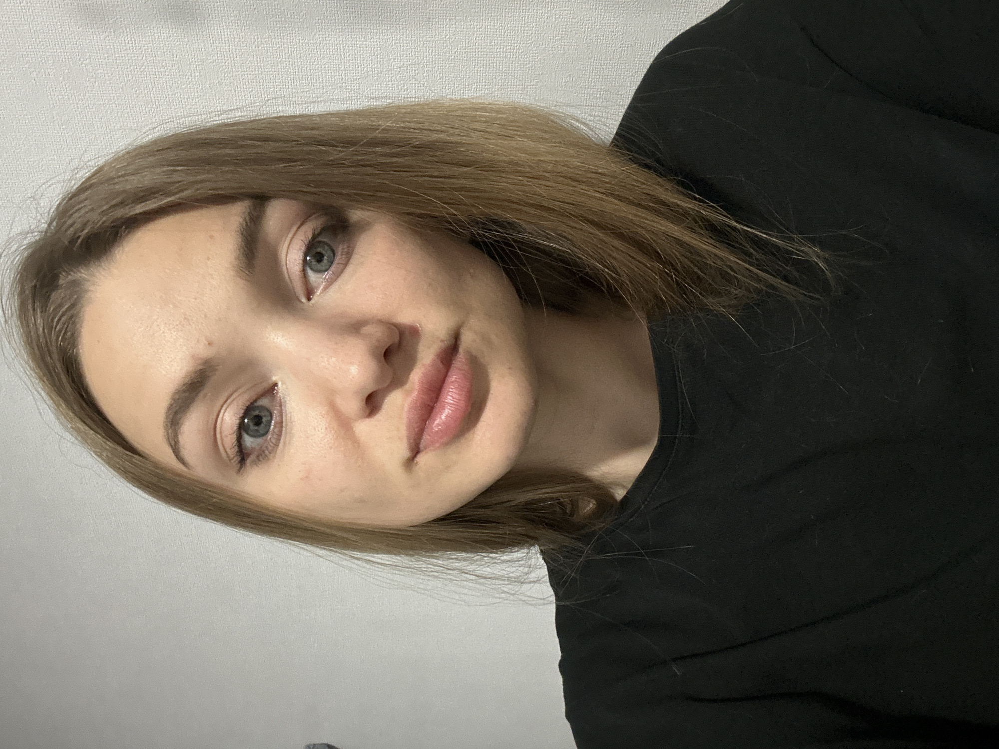 A close-up, slightly angled selfie of a young woman with fair skin and light brown hair. She is wearing a black top. Her eyes are light blue or grey. The background is a plain, textured white wall. There is no indication of the location being Kazan, Russia.
