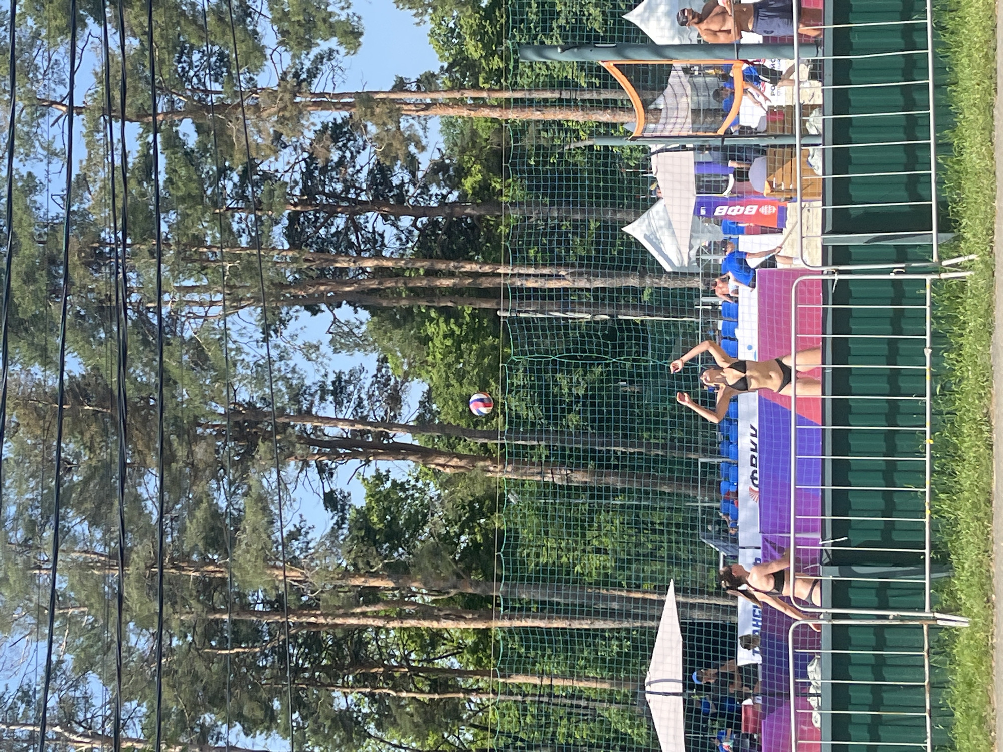 The image depicts a beach volleyball game in what appears to be a park setting, near Krasnodar, Russia. Two female players in black bikinis are actively engaged in a volley, with one player reaching to hit the ball mid-air. A net divides the court. Behind the court are seated spectators, and beyond them are tall trees, likely pines. Along the perimeter are barriers. The location is possibly in or near Krasnodar, Russia, based on the Cyrillic lettering present on some of the signage in the background.