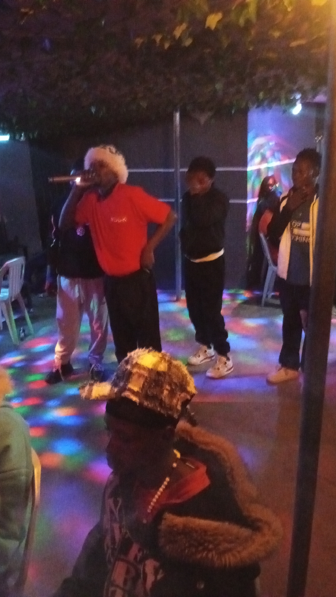 The image captures an indoor scene in the city of Unknown, Unknown, characterized by dim ambient lighting and multi-colored disco light patterns illuminating the floor. Overhead, dark foliage is visible.
There are five main individuals present, with additional indistinct figures in the background. In the center-left, a male individual with dark skin is positioned, wearing a white fluffy hat, a red t-shirt with "YOGK" printed on the chest, and dark pants. He holds a metallic microphone to his mouth with his left hand, while his right hand rests on his hip. To his left, another male individual with dark skin wears a black top, light-colored pants, and dark shoes, standing slightly behind the central figure.
To the right of the central figure, a male individual with dark skin wears a black long-sleeved top and black pants, paired with white sneakers. He stands with his arms appearing to be crossed or his hands tucked into pockets, looking downwards. To his right, another male individual with dark skin wears a blue t-shirt with white text including "TOP THING," a white and black jacket, and light-colored sneakers. He is facing slightly left.
In the foreground, positioned at the bottom center and slightly out of focus, a male individual with dark skin is visible, wearing a multi-textured, possibly camouflage, cap. He has on a dark jacket with a brown fur-trimmed collar and a red/orange shirt underneath, accessorized with a light-colored beaded necklace. His head is turned slightly to the left.
The environment includes structural elements such as dark vertical posts and horizontal bars in the background. A white plastic chair is partially visible on the far left. The floor exhibits distinct patches of red, green, blue, and yellow light.