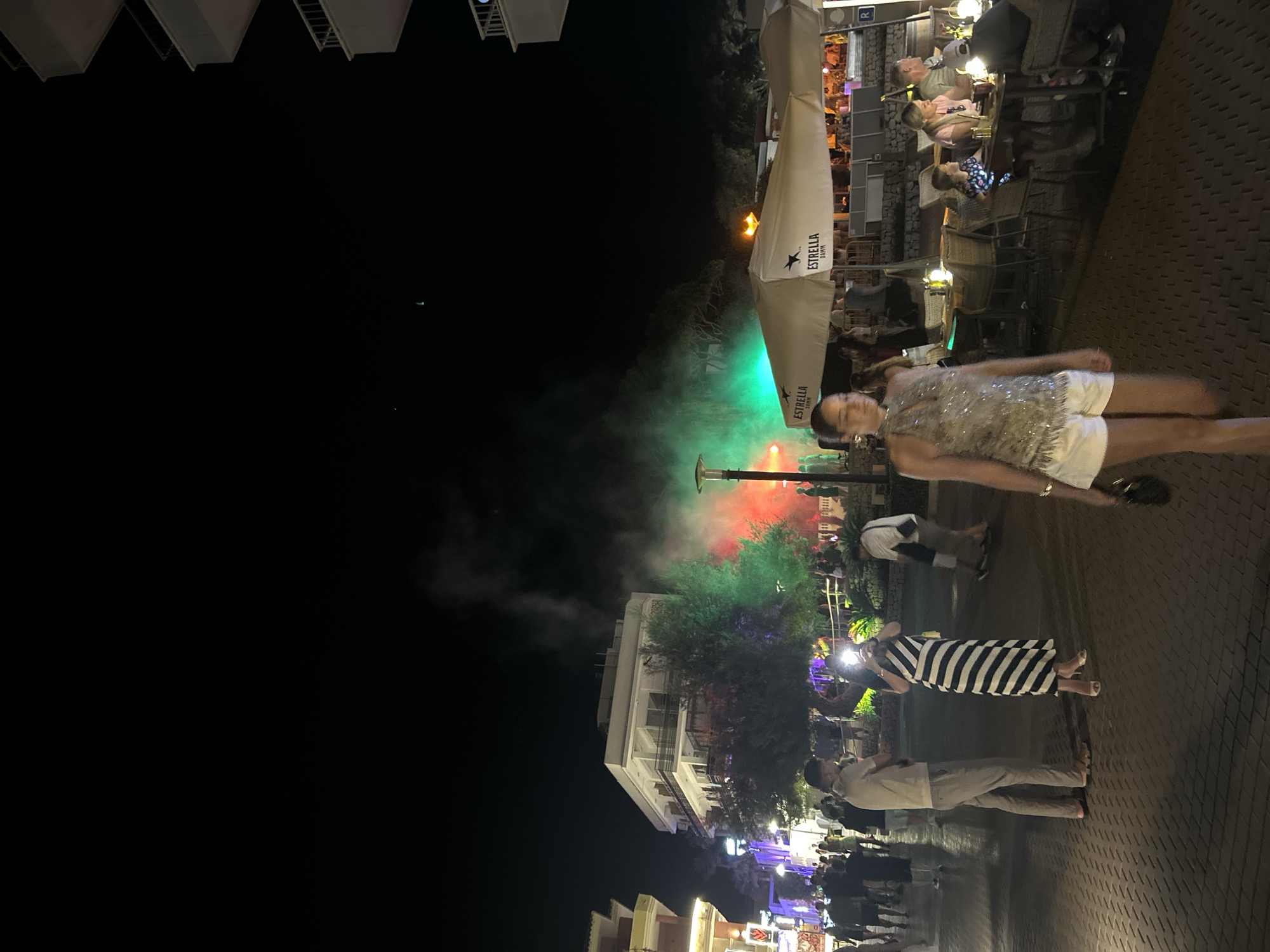 The image depicts a night scene in Cala Rajada, Spain, likely on a pedestrianized street lined with buildings and outdoor seating areas for businesses. The foreground shows a paved walkway with several people walking and standing. A woman in a sparkly halter top and white shorts is walking towards the camera. Further back, a woman in a black and white striped dress is taking a photo with her phone. A man stands next to her.
In the background, there is a building with multiple balconies. To the right of the building, a large area with outdoor seating and umbrellas is visible, suggesting a restaurant or bar. One umbrella prominently displays the "Estrella Damm" logo. Further back, there is a spectacle of colored smoke or fog, illuminated by red and green lights, creating a vibrant and atmospheric effect. Trees are visible amidst the smoke. The overall scene suggests a lively evening in a tourist area.