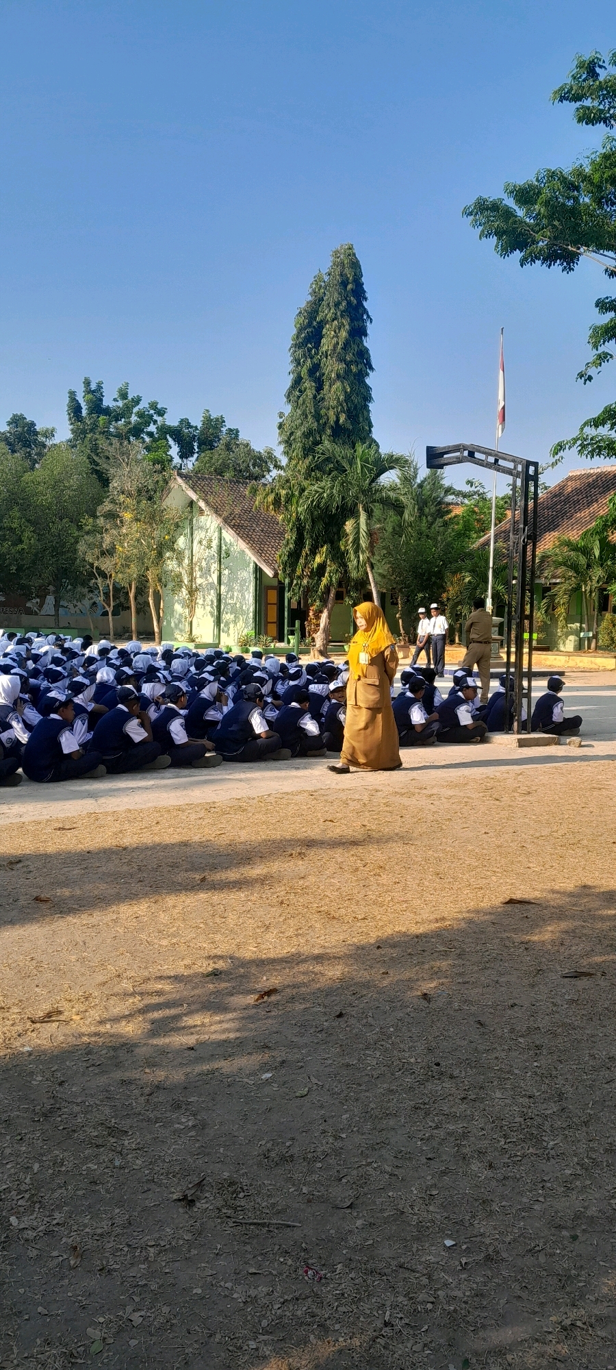 In Tahunan, Indonesia, a group of students, likely in uniform, are seated on the ground in an outdoor area. A woman in a tan hijab and matching attire stands to the right of the group, appearing to address them. In the background, a light green building with a brown roof is visible, surrounded by trees. A flagpole with a white and red flag stands near the building. The ground is a mixture of dirt and dry leaves. The scene suggests an outdoor assembly or class taking place at a school.