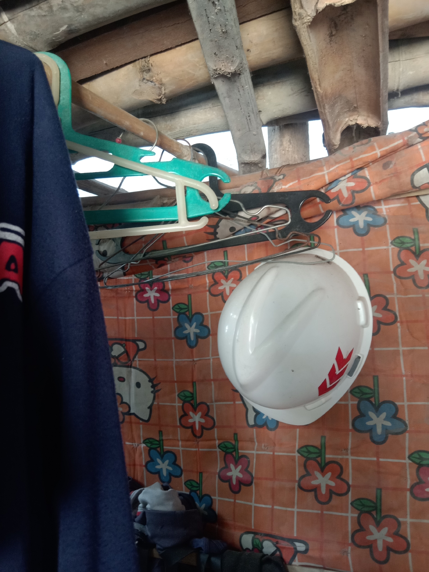In Stiaji, Indonesia, a white hard hat with red accents rests on an orange cloth patterned with flowers and cartoon characters. Several hangers are visible, some holding clothing, and a dark blue garment hangs to the left of the frame. The scene appears to be outdoors or in an open-air structure, with wooden beams and a rustic roof overhead.
